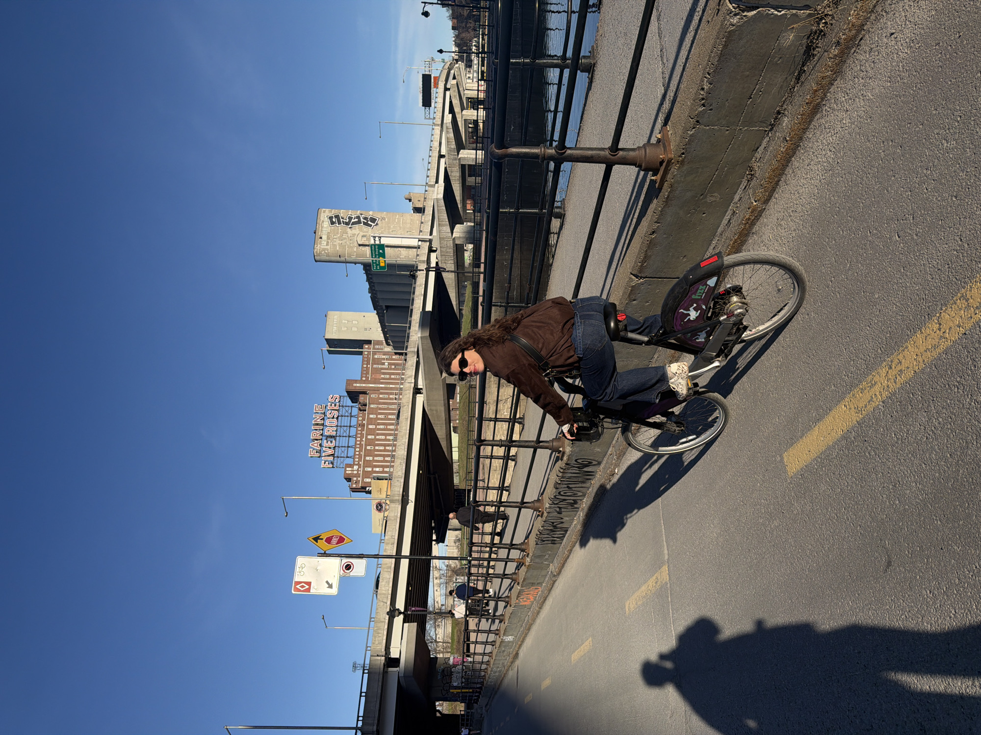 A woman wearing a brown jacket, dark jeans, and sunglasses rides a bicycle on a paved path next to a concrete barrier with a metal railing. The path is marked with yellow lines. In the background, there are buildings, including a grain elevator covered in graffiti, and the "Farine Five Roses" sign, indicating this is Montreal, Canada. There are also street signs and utility poles. The sky is blue and the sun is shining, casting a shadow of the photographer on the path. The body of water behind the barrier would most likely be the St. Lawrence River.