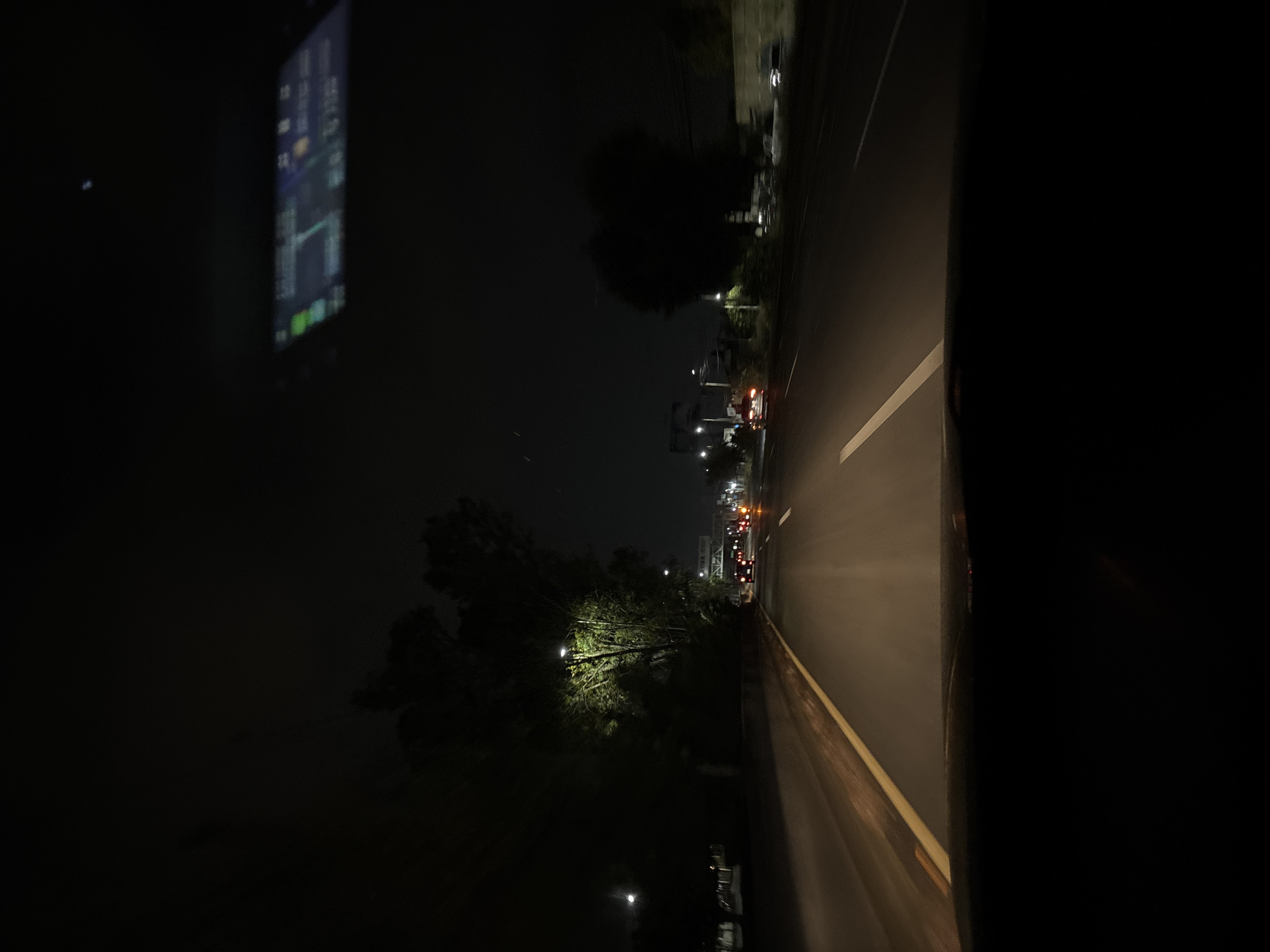 A nighttime, low-angle shot from inside a vehicle traveling on a road in what appears to be a city. The view is of the road ahead, illuminated by streetlights and the vehicle's headlights. A yellow line marks the road edge. There are other vehicles ahead, with visible tail lights. Trees and buildings line the road. A screen, possibly a navigation device or phone, is partially visible in the top left of the frame. The location is the city of Puebla, Mexico.