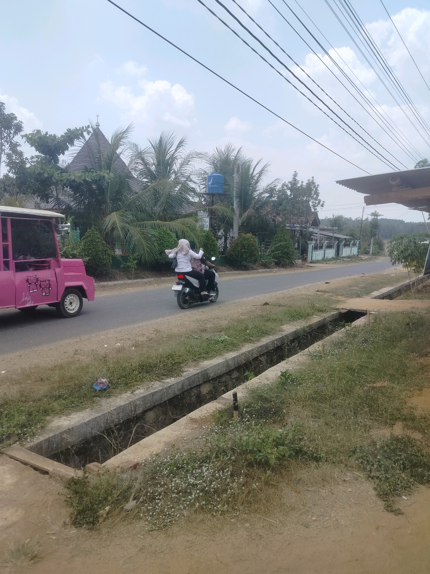In the city of Rejosari, Indonesia, an outdoor scene unfolds along a paved road. To the left, a bright pink, open-sided vehicle resembling a golf cart, adorned with Hello Kitty decals, is parked on the unpaved shoulder. The vehicle has a white roof and a white circular wheel visible. A motorcycle is traveling on the road, carrying two individuals. The person driving wears dark clothing and a helmet. The passenger in the back, a woman, is wearing a light-colored hijab and a long-sleeved shirt, with her arms raised. The road continues into the distance, bordered by a concrete drainage ditch on the right. The ditch is partially filled with water and surrounded by overgrown grass and small white flowers. Beyond the road, to the left, a distinctive building with a dark, angled roof topped with a crescent moon symbol is visible amongst palm trees and other foliage. Various houses and structures are situated further back, with power lines stretching across the sky. The sky is overcast with scattered clouds.