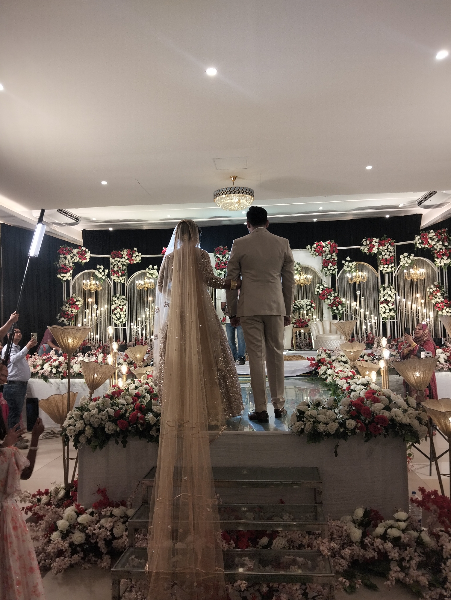 The image shows a wedding ceremony taking place indoors, likely in a city called Unknown, in an area called Unknown. The focus is on a bride and groom standing on a slightly elevated platform. The bride is wearing a long, ornate dress with a long veil that extends down the stairs leading to the platform. The groom is in a light-colored suit. Both are facing away from the camera, towards a decorated backdrop.
The backdrop consists of a black curtain interspersed with white archways decorated with red and white flowers, as well as chandelier-style lights. The platform itself is also decorated with an abundance of flowers in similar colors. Below the platform, more flowers are arranged, cascading down the stairs.
To the sides, there are several attendees, some of whom are taking pictures with their phones. There is a light stand on the left side of the image, suggesting professional photography or videography is present. The room is well-lit, with ceiling lights visible.