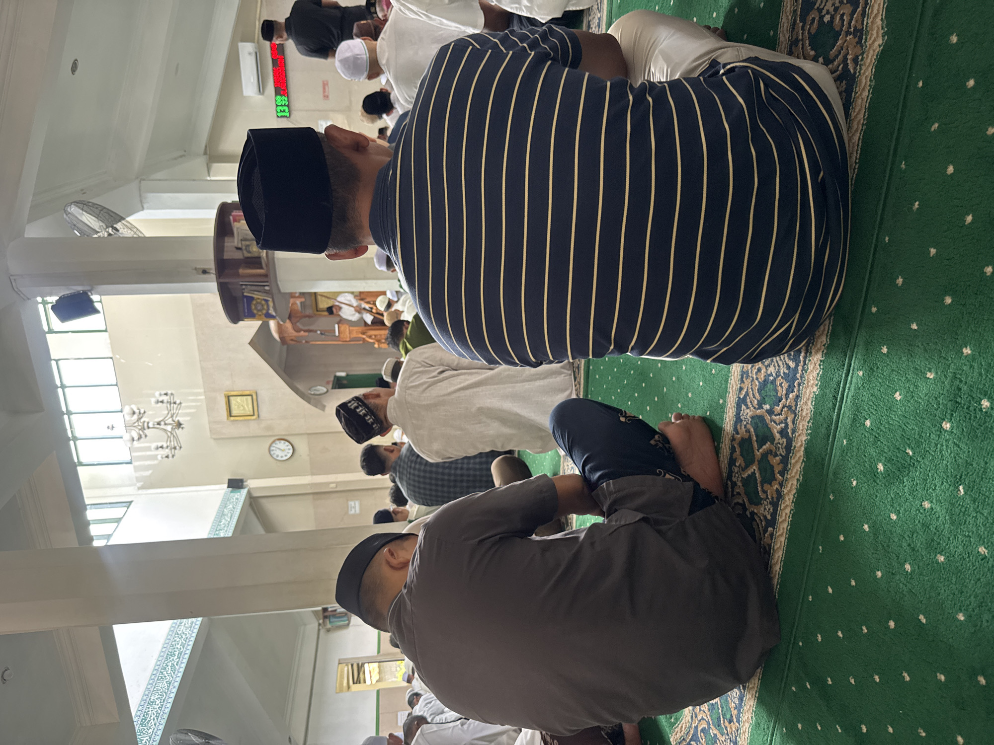 The image shows the interior of a mosque in Banda Aceh, Indonesia, where several men are seated on a green carpeted floor facing the same direction, presumably for prayer. Two men are prominently featured in the foreground, their backs to the camera. The man on the left wears a dark gray short-sleeved shirt and dark pants, with a black peci on his head. The man on the right wears a navy blue and yellow striped shirt, light-colored pants, and a black peci. Other men in various attire, including white thobes and pecis, are visible in the background. The mosque features high ceilings with decorative elements, chandeliers, and large windows. A clock is visible on the wall in the background, along with an air conditioning unit and a digital display. The overall atmosphere suggests a moment of communal prayer or religious gathering.