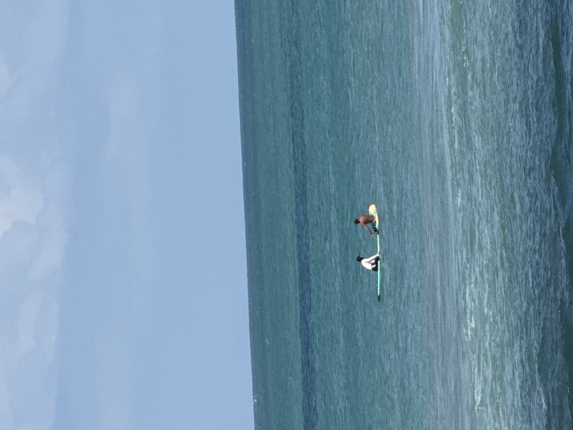 Two individuals are on paddleboards in the ocean. One person, wearing a white shirt and dark shorts, is seated on a green paddleboard. The second person, shirtless with dark shorts, is seated on a yellow paddleboard. They are facing each other and appear to be interacting. The water is a deep blue-green, with some whitecaps visible in the foreground, suggesting moderate waves. The horizon line divides the ocean from a pale blue sky with some wispy white clouds. The location is Kuta, Indonesia.