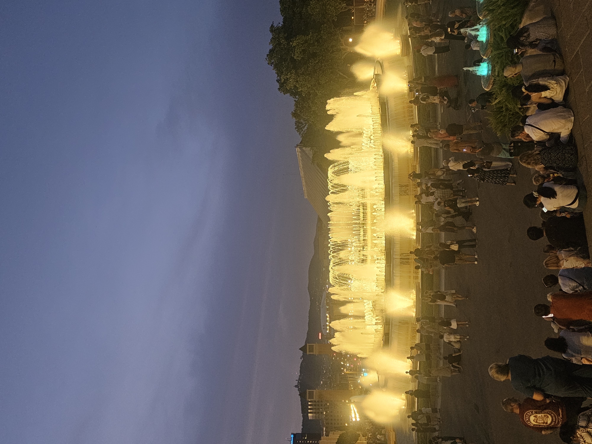 The image depicts the Magic Fountain of Montjuïc in Barcelona, Spain, at dusk. A large crowd of people are gathered, seated and standing, watching the illuminated fountain which is spewing jets of water that glow with a warm, yellow light. In the background, the city skyline is visible, with buildings and lights extending into the distance. The sky is a deep blue, transitioning to purple, with some clouds. The overall scene conveys a sense of public gathering and enjoyment of a light and water spectacle.