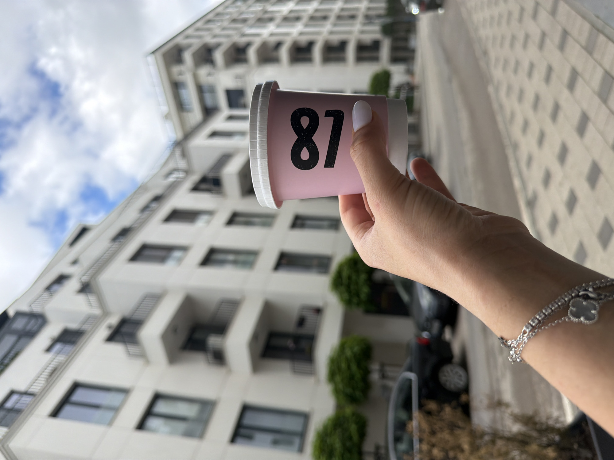The image shows a human hand holding a light pink or purple cup with the number "87" printed in black on its side. The hand, likely female, has a thumb with white nail polish and wears a silver bracelet adorned with a four-leaf clover charm and a second chain featuring small, possibly gem-like, embellishments.
The background reveals a tall, modern building with a light-colored facade, numerous windows, and structured balconies, indicating an urban setting. Below the building, a paved area, likely a sidewalk or street, is visible with a geometric block pattern. Green shrubbery can be seen near the building's foundation, and parts of parked vehicles are faintly discernible. The sky above is partly cloudy with patches of blue, suggesting a daytime setting. The lighting creates distinct shadows on the paved ground, consistent with morning or afternoon light. The scene is set in an urban environment in Sofia, Bulgaria.