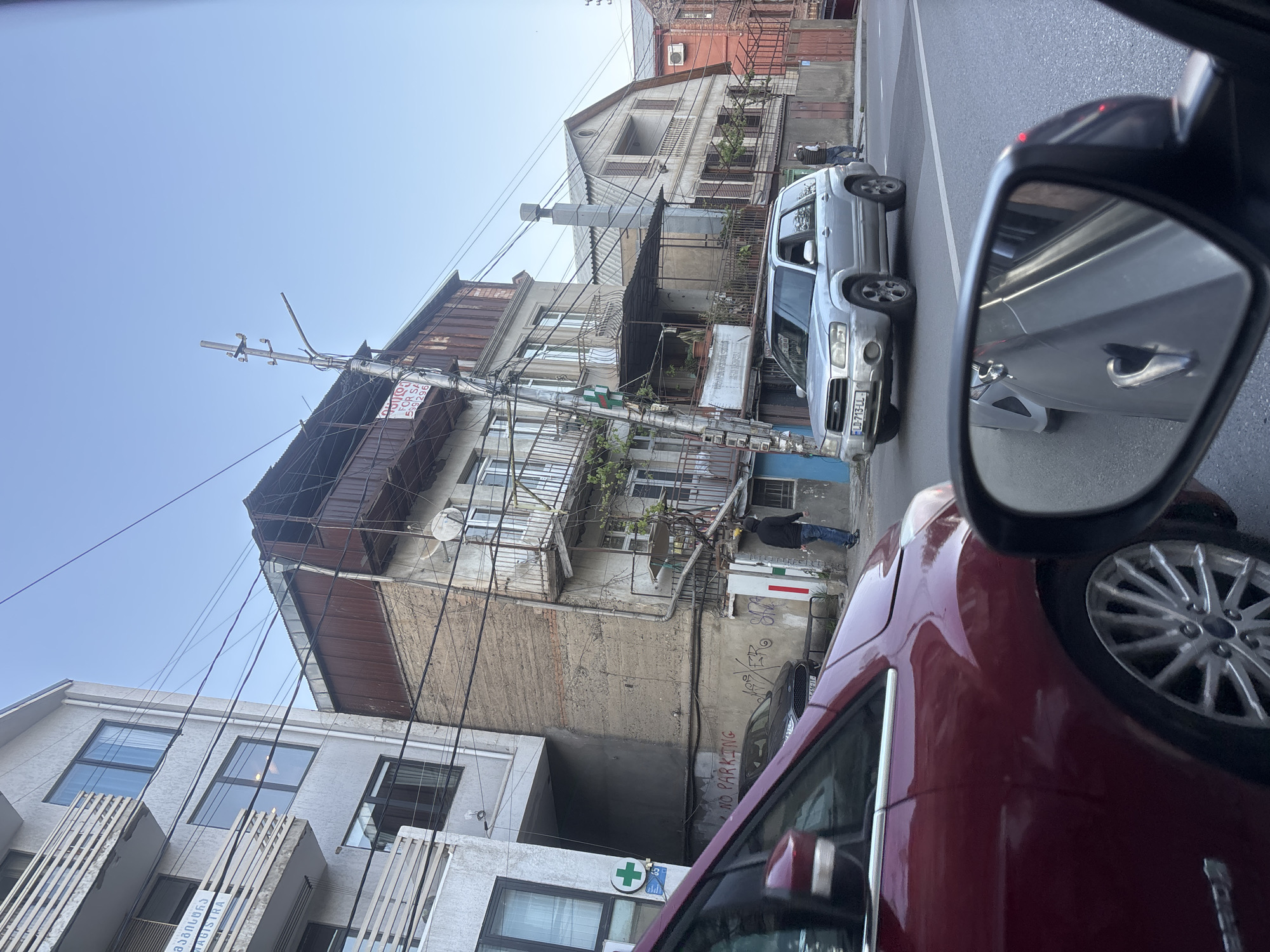 This media file captures an urban street scene in Tbilisi, Georgia, viewed from the perspective of a vehicle, with a prominent side mirror on the right. The image is taken in bright daylight under a clear blue sky.
The scene features several multi-story buildings of varying architectural styles. On the left, a more modern-looking building with balconies and large windows is partially visible. Opposite it, a older building features weathered concrete, numerous crowded balconies, and extensive, tangled utility wires and cables running vertically and horizontally. This building has a "FOR SALE" sign in Georgian and English ("Colle FOR SA 599 296"). Further down the street, another building with a distinctive roof and more traditional balconies can be seen.
In the foreground, parts of a dark red car are visible, including its side panel, tire, and chrome trim. A silver Subaru Forester SUV with a license plate "ELD-713-LL" is parked or stopped next to the red car. Its reflection, along with another vehicle, is visible in the side mirror. A person wearing a dark jacket and blue pants is standing near the entrance of one of the buildings.
Visible text includes the aforementioned "FOR SALE" sign, and another white sign with a green cross symbol and the text "მაგისხია MAGISTRA", indicating a pharmacy or medical establishment. On a concrete wall, "NO PARKING" is spray-painted in red. The overall impression is a bustling urban environment with a mix of old and new architecture and dense infrastructure.