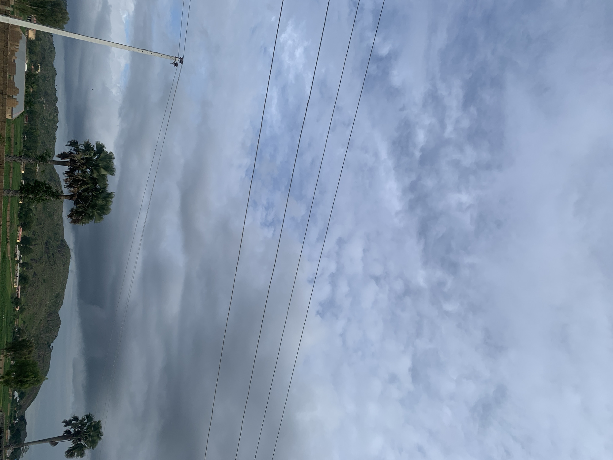 The image depicts a cloudy sky over Kuru Karama, Nigeria. Several power lines are visible stretching across the frame. In the foreground, a utility pole stands to the left. Two palm trees are prominent in the mid-ground, with a third palm tree further to the right. A hilly landscape with scattered buildings and green fields forms the background. The overall impression is of a rural or semi-rural environment under an overcast sky.