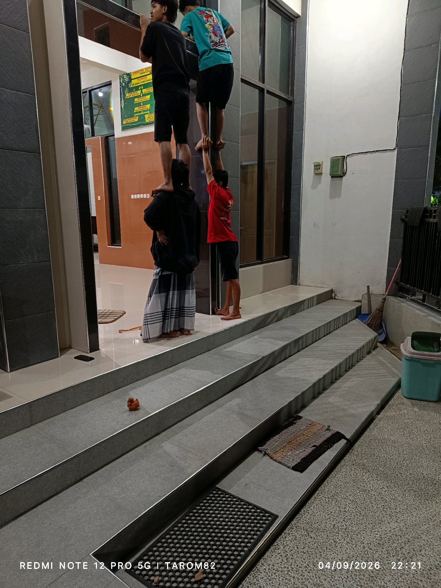 The image depicts four young males in what appears to be an outdoor, covered walkway or entrance area with a set of stairs leading down. The location is identified by the timestamp as being in Rejoagung, Indonesia.
The individuals are arranged in a human pyramid formation on a raised platform or the first step of the stairs.
The bottom-most person is obscured, with only their lower body and feet visible, wearing a sarong.
The second person from the bottom is wearing a black hoodie and black shorts and is standing on the shoulders of the person below.
The third person from the bottom is wearing a teal t-shirt with graphics and black shorts, standing on the shoulders of the person in the hoodie.
The top-most person is wearing a red t-shirt and dark shorts, and appears to be balancing on the hands of the person in the teal t-shirt.
The environment consists of tiled floors and walls, with a dark, grey textured wall on the left and a white wall on the right. There are large glass doors or windows visible in the background, leading into a well-lit interior space. The stairs are made of grey concrete or stone with metal edging. On the stairs, there are two small mats, one with a woven pattern and another black, rubber mat with a circular pattern. A small, orange-brown object, possibly a piece of debris or a decorative item, is also on one of the steps. A broom and dustpan are leaning against the wall near the bottom of the stairs, and a turquoise plastic bin is situated to the right of the stairs. The lighting suggests it is evening or nighttime. The timestamp in the bottom right corner indicates the image was taken on 04/09/2026 at 22:21.