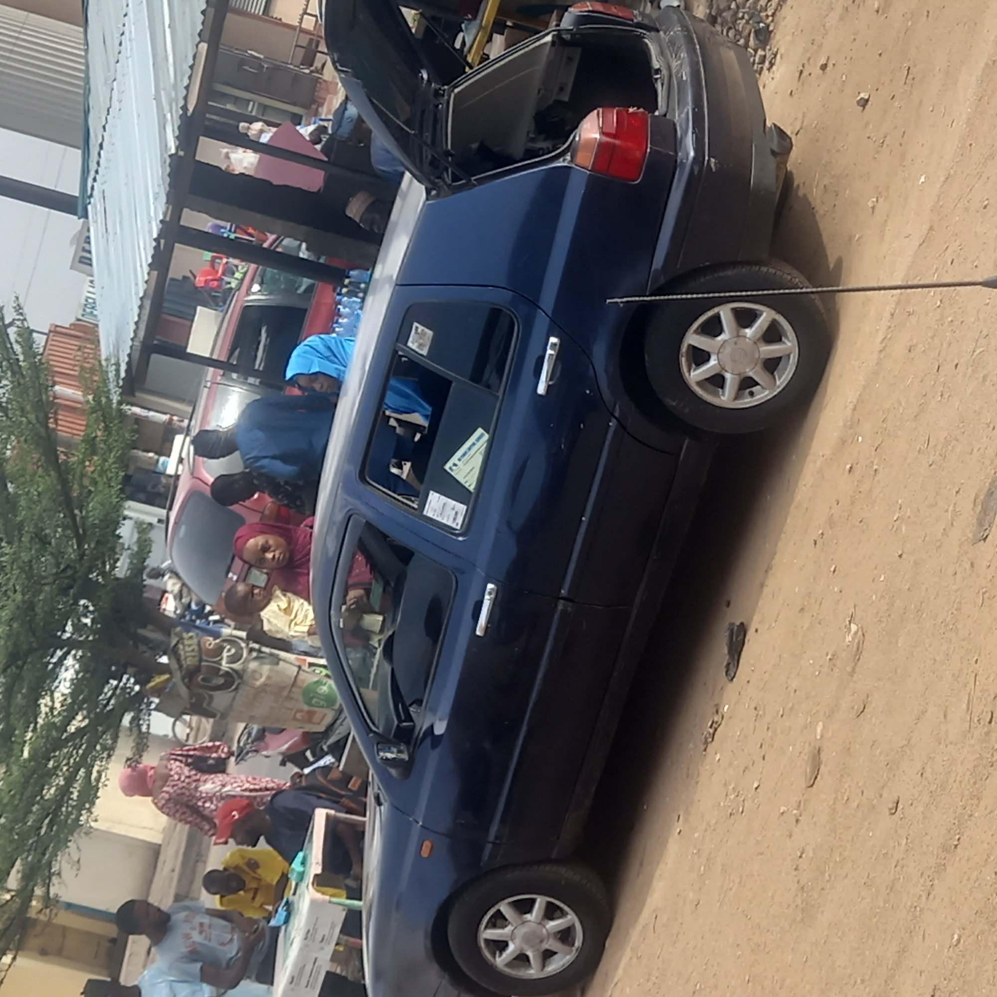 The image depicts a dark blue hatchback-style car parked on a dusty lot in Dadi Riba, Nigeria. The car's trunk is open, and there are several people gathered around the vehicle and a table set up nearby, possibly engaged in some sort of transaction. The surroundings include buildings with corrugated metal roofs, a tree, and other vehicles. The scene appears to be a casual outdoor gathering, likely related to commerce or community activity.