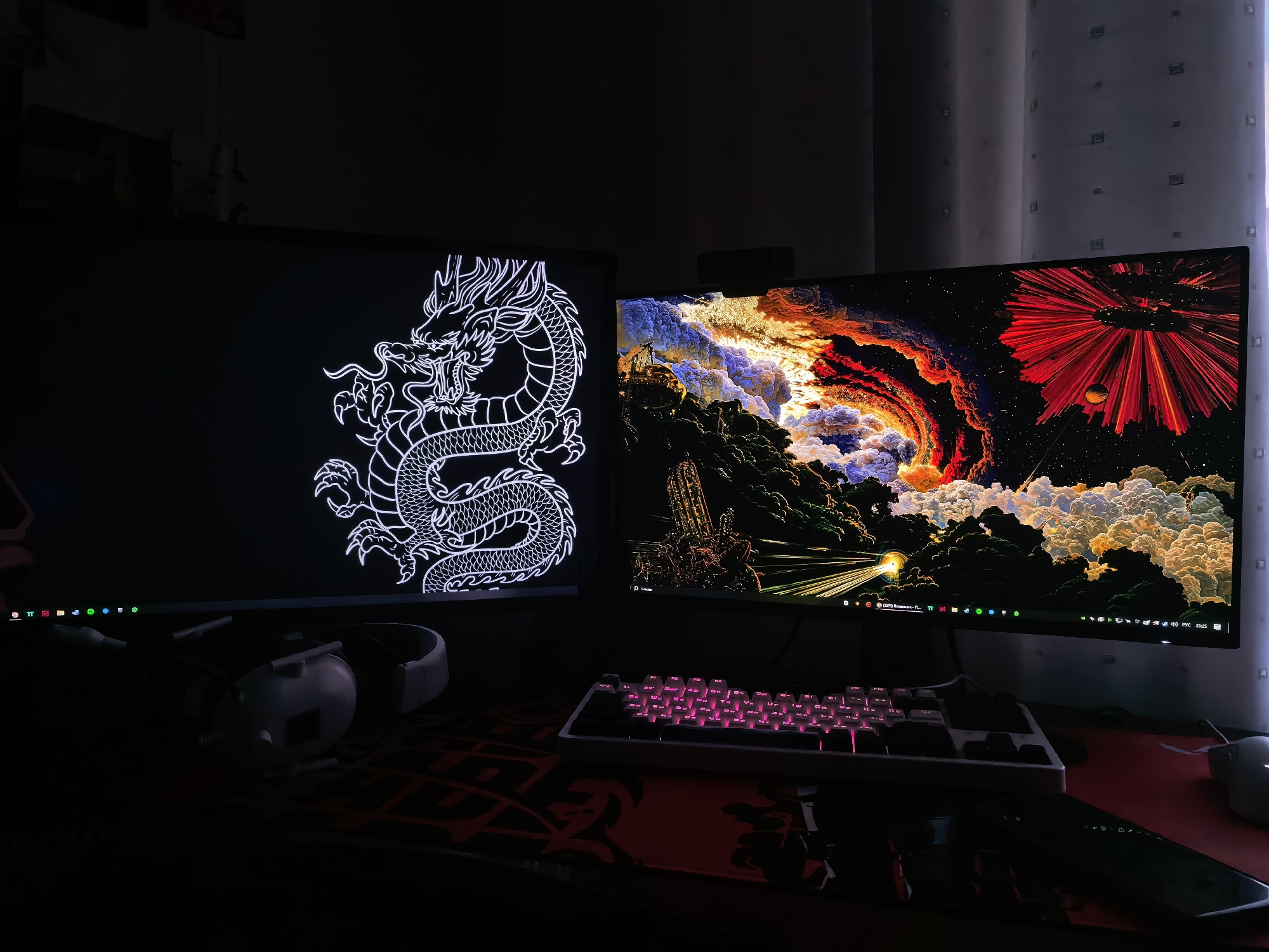 The image shows a desk setup with two computer monitors, a keyboard, and headphones. The left monitor displays a white outline of a dragon on a black background. The right monitor shows a vibrant, abstract image with swirling clouds in shades of orange, red, and blue, and a red, spiky object in the upper right corner. A white keyboard with pink backlighting is in front of the right monitor. A pair of white headphones rests on the desk to the left of the keyboard. The desk itself appears to be covered by a red mousepad with a black pattern. The scene is dimly lit, with the light primarily coming from the monitors and the keyboard backlighting. The background consists of a dark wall and sheer curtains. The location is unknown, and no people are visible.