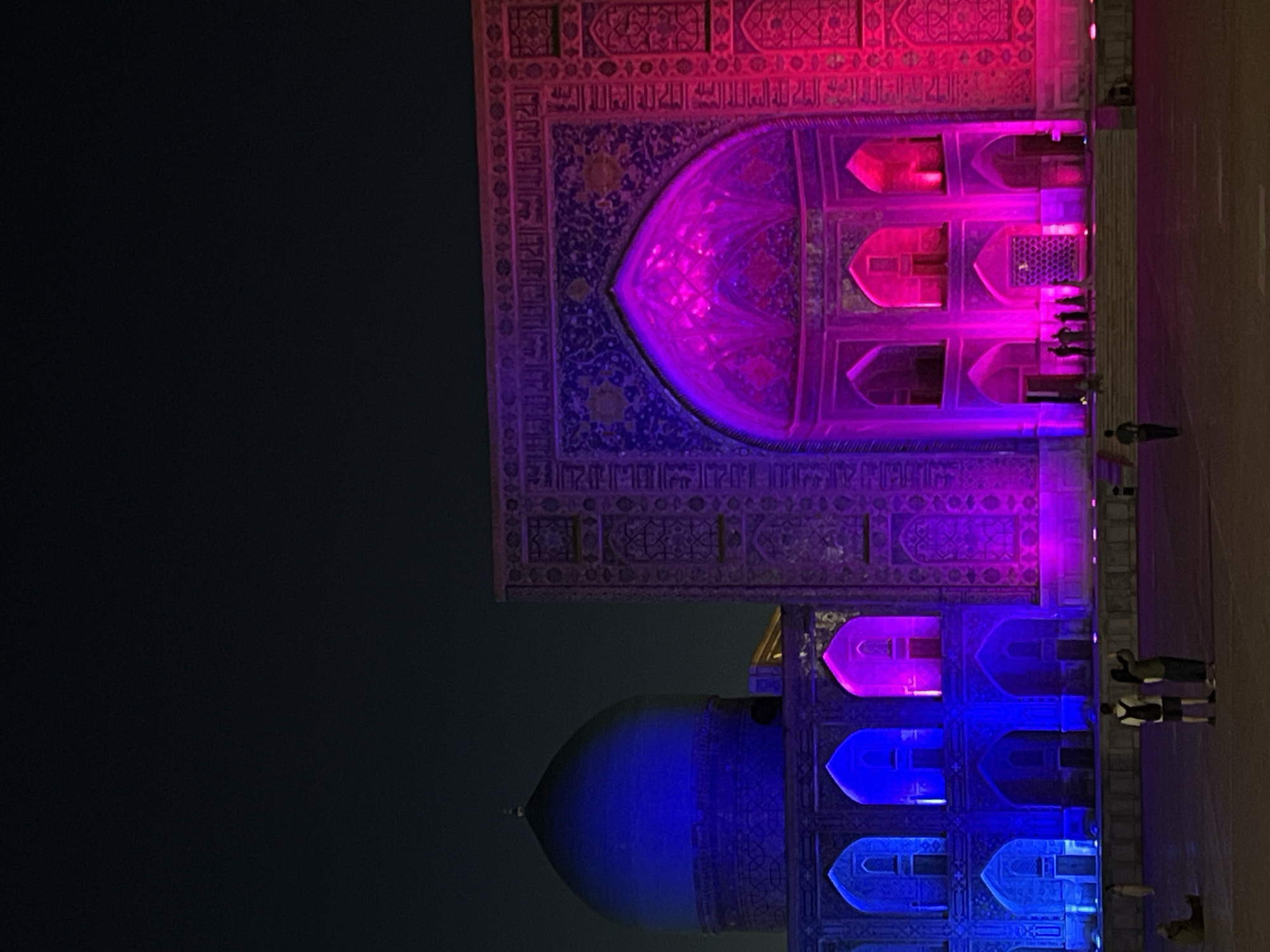 The image captures a nighttime scene of an ornate building illuminated by vibrant colored lights. The architecture is intricate, featuring decorative carvings and arched windows, characteristic of historical Middle Eastern or Central Asian structures. The building is bathed in a gradient of colors, transitioning from deep blue at the bottom to shades of purple and pink towards the top, with some areas highlighted in bright magenta.
Several small figures of people are visible along a pathway or plaza in front of the illuminated structure, suggesting a public space. One person is walking away from the camera on the right side of the image, while a group of silhouetted figures stands near the base of the building. Their presence provides a sense of scale and indicates that the scene is accessible to visitors.
The lighting creates a dramatic and almost ethereal atmosphere, emphasizing the detailed ornamentation of the facade. The time of day is clearly night, given the dark sky and the artificial illumination of the building. There are no explicit weather cues, but the darkness suggests clear conditions. The architectural style strongly suggests the location is Samarkand, Uzbekistan, known for its historical Islamic architecture. No visible text is present in the image. The overall impression is one of awe and wonder, highlighting the beauty of the historical site through a modern light display.