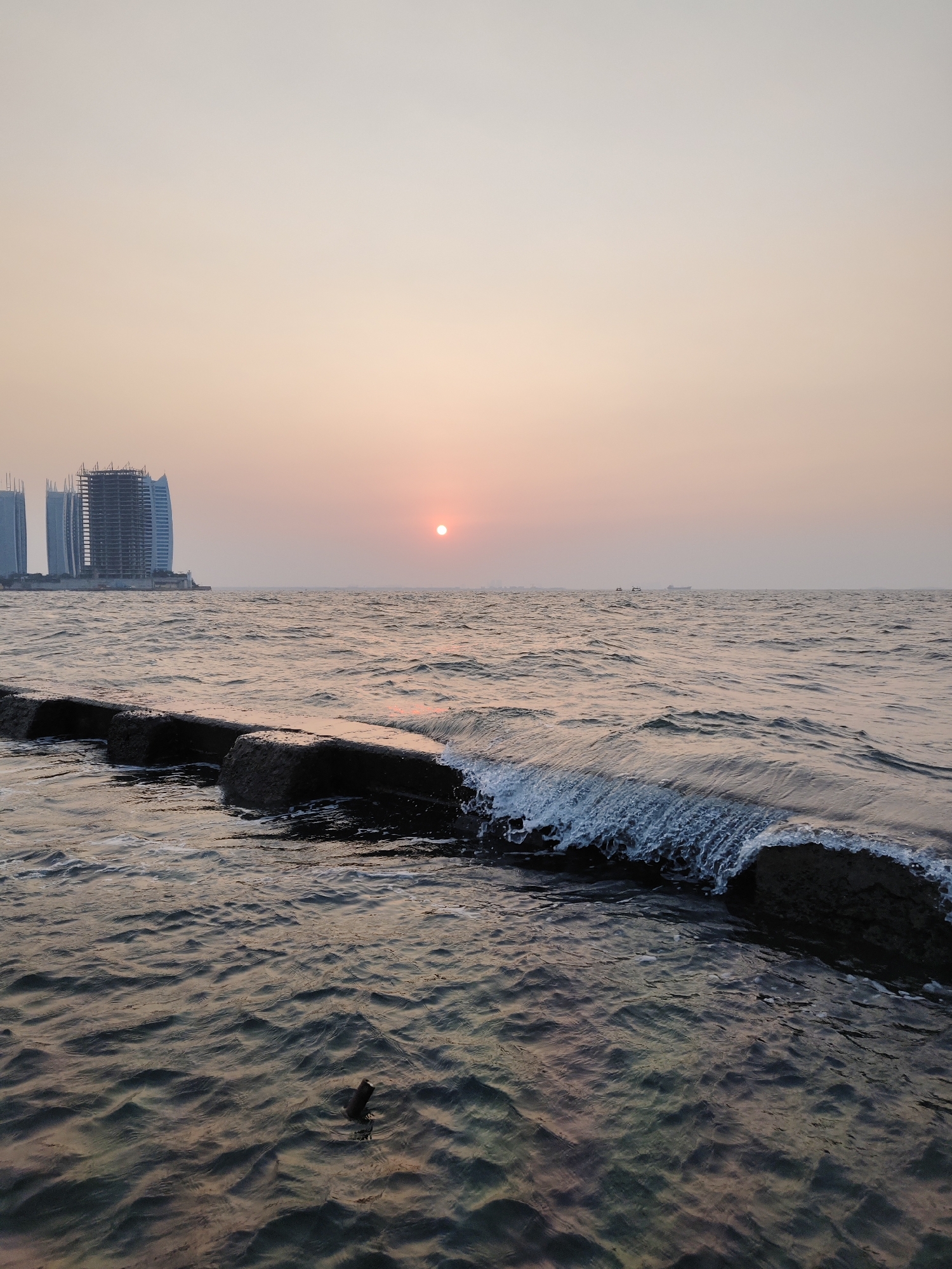 The image depicts a sunset over the ocean, with a line of large, dark rocks extending into the water in the foreground. Waves are breaking against the rocks, creating white foam. In the background, there are several tall buildings, some of which appear to be under construction. A small, dark object, possibly a piece of debris or a small bird, is visible in the water to the right of the rocks. The sky is a gradient of soft pinks and oranges, fading into a pale yellow towards the top. The overall atmosphere is calm and serene, with the setting sun casting a warm glow over the scene.