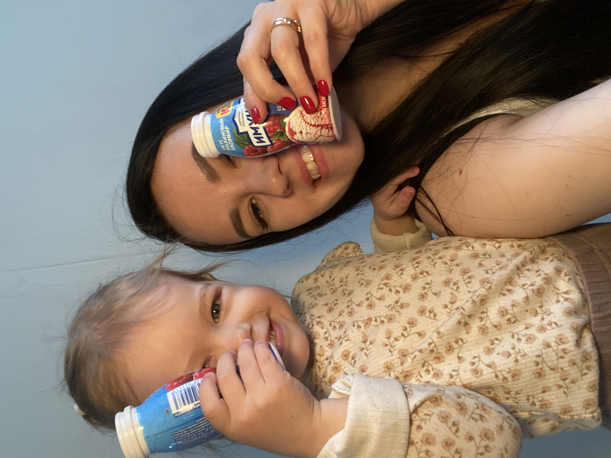 The image depicts a woman and a young child posing for a photo indoors. The woman has long, dark hair and is wearing a ring on her ring finger. She is holding a small bottle of what appears to be a yogurt drink, placing it near her face. The child, presumably her daughter, also has a similar bottle of the same drink in her hands. The child is wearing a floral-patterned top. The background is a light blue wall. The brand name of the drink is "Imun". The location is Chaikovsky, Russia.