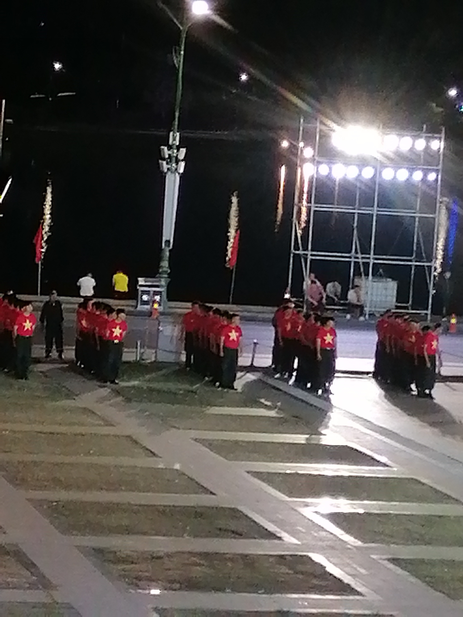 The image depicts an outdoor nighttime scene, likely in Da Lat, Vietnam, judging by the presence of fireworks and a formal gathering. A group of individuals, predominantly young men, are lined up in formation, wearing matching red shirts with yellow stars and dark pants. They appear to be participating in a coordinated event or performance. In the background, a scaffolding structure with multiple bright lights is visible, suggesting a stage or illuminated backdrop. There are also several people standing further back, including two in casual attire. The ground is a paved area with grassy sections, separated by pathways. A tall lamppost with multiple light fixtures stands prominently on the left side of the image. The overall atmosphere suggests a celebratory or official occasion.