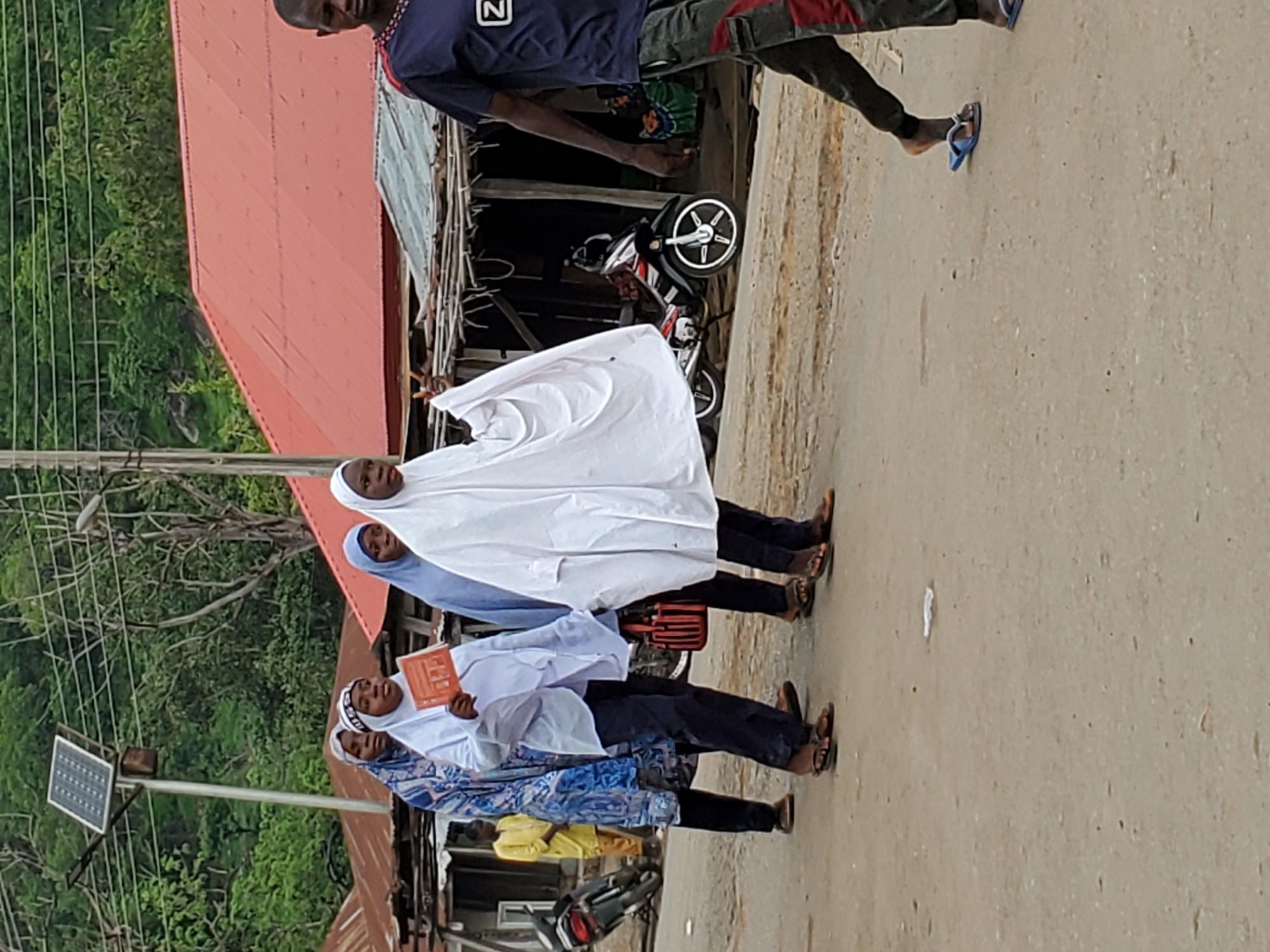 In Mararaba, Nigeria, three young women wearing hijabs and carrying books walk along a paved road. One woman is covered in a white hijab, another in light blue, and the third in a blue patterned one. A man wearing a dark blue shirt and dark pants walks away from the women in the opposite direction. To the left of the women is a solar panel attached to a utility pole. Adjacent to the road is a structure with a red roof. Behind the women is a parked motorcycle.