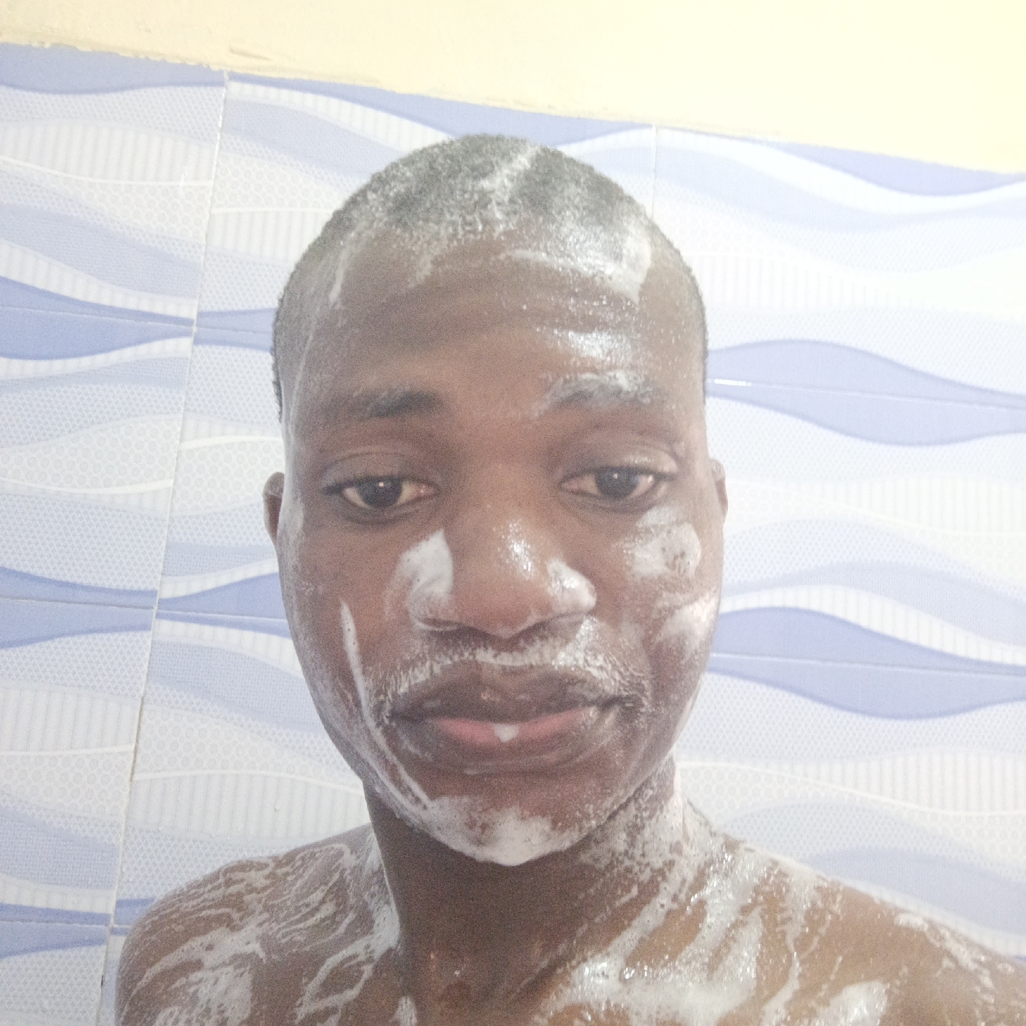 A close-up shot shows a dark-skinned individual, likely male, with short hair, covered in white soap or foam. The foam covers his face, head, neck, and upper chest. The background consists of blue and white patterned tiles. Based on the prompt, the location is assumed to be Keffi, Nigeria.