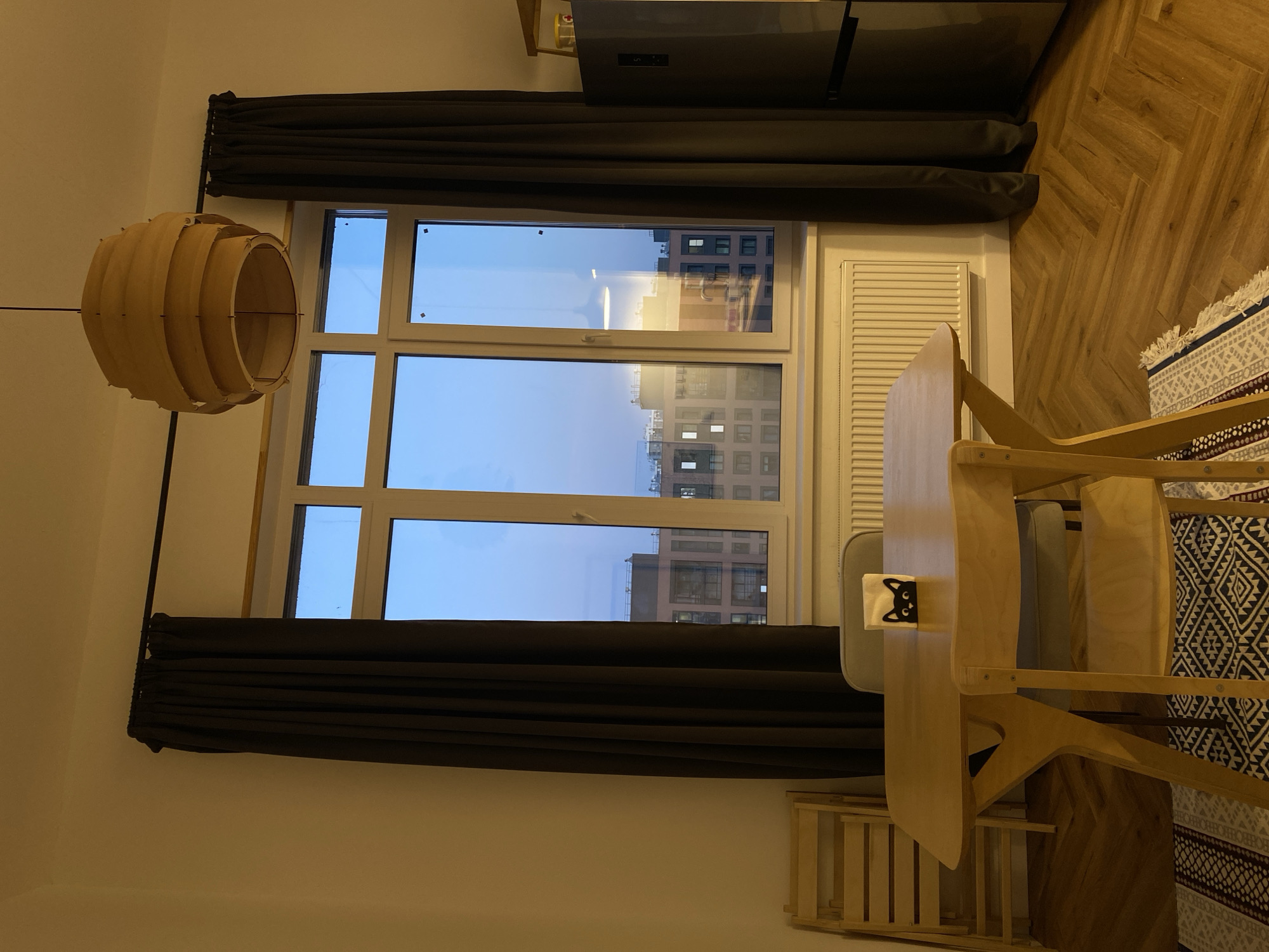 Here's a description of the image:
The image shows an interior scene. The scene is primarily focused on a window with dark curtains on either side. Outside the window, buildings are visible, reflecting the early evening or late afternoon light, possibly from Izhevsk, Russia.
A wooden table and chair set are positioned to the right of the window. A light fixture, also made of wood, hangs to the left of the window. A radiator is present on the wall below the window. Part of a grey refrigerator can be seen in the upper part of the image. The floor is made up of light-colored wood planks arranged in a herringbone pattern. A rug with a patterned design is partially visible at the edge of the frame near the table. There are no people visible in the image.