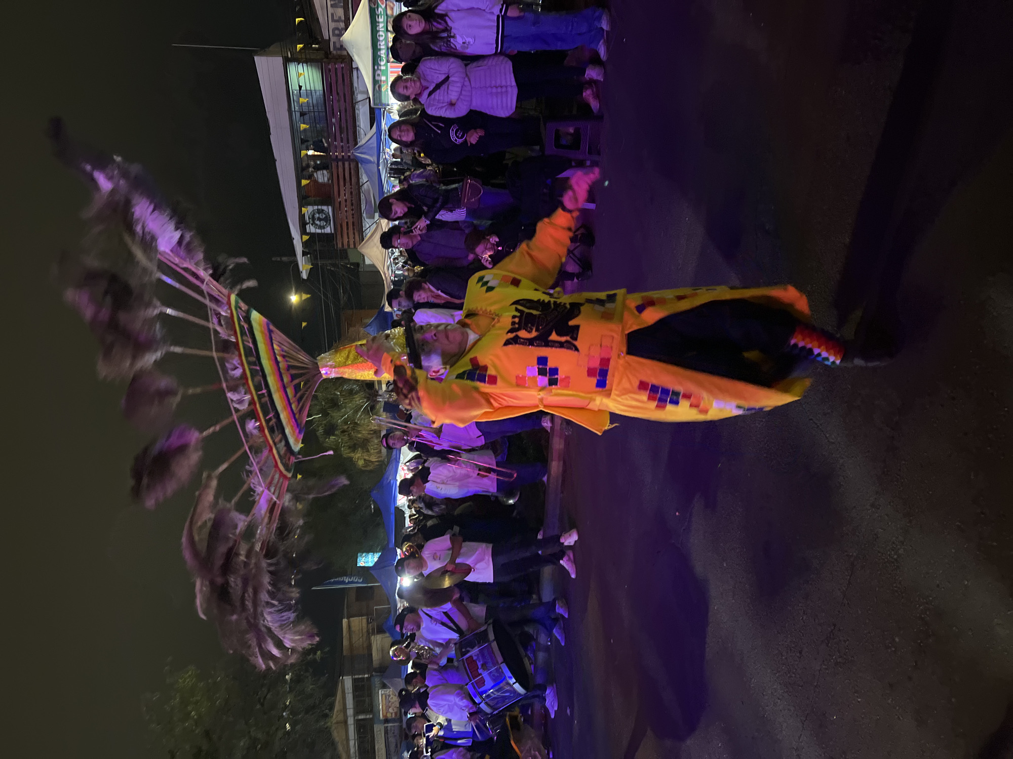 In Arica, Chile, a street festival is underway at night. A performer in a vibrant yellow costume with a feathered headdress is the central focus, captured mid-dance or movement. The headdress, adorned with colorful fabric strips and long, dark feathers, is dramatically swept upwards and to the left, blurring due to motion. The performer's outfit is elaborately decorated with square patterns of red, blue, and yellow, and features a crest on the chest. They are on a paved street illuminated by purple and white lights, casting long shadows.
Behind the performer, a lively crowd is visible. A brass band is playing, with members holding drums, cymbals, and trombones. They are dressed in white shirts and black caps. Spectators stand on the side, some looking towards the performer, others engaged in conversation or observing the festivities. In the background, tents and buildings suggest a market or fair setting, with signage including "PICARONES" visible on one tent. The overall atmosphere is festive and energetic, typical of a cultural celebration in a city.