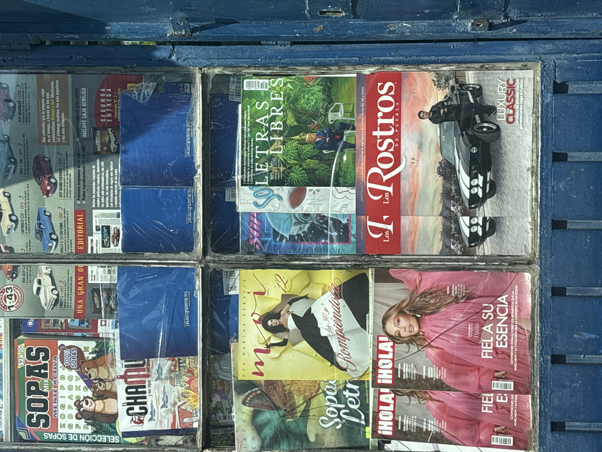 The image shows a magazine rack filled with various magazines. The magazines include "Letras Libres," "Los Rostros de Puebla," and "¡Hola!". The cover of "Letras Libres" features a person sitting in a chair in a garden. "Los Rostros de Puebla" has a picture of a man standing next to a car. "¡Hola!" features the celebrity Thalia. Other magazines are stacked below with puzzles on their covers. The location is Puebla, Mexico, as indicated on the magazine "Los Rostros de Puebla." The magazine rack appears to be made of a blue-painted metal.