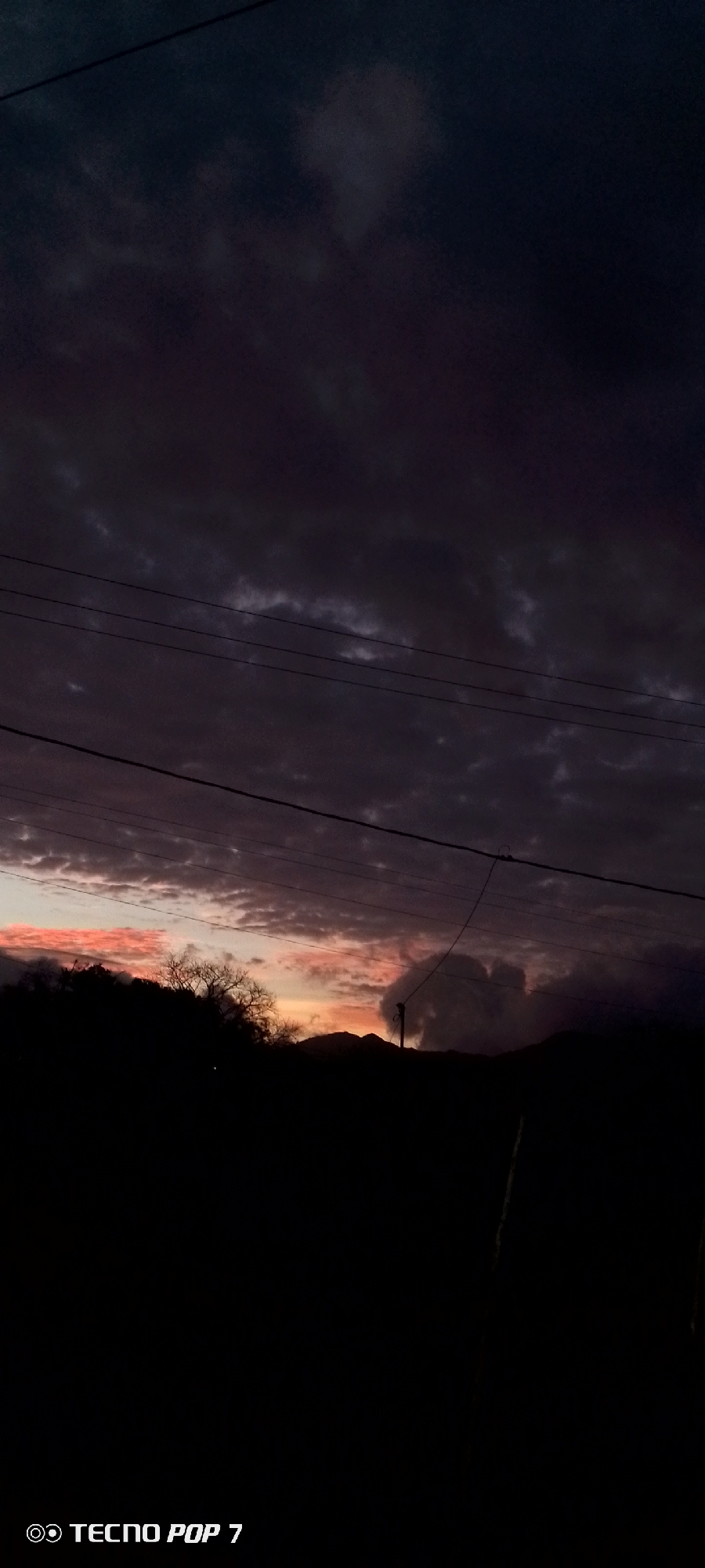 The image depicts a dark, overcast sky at sunset in Altagracia, Venezuela. The horizon is illuminated with a reddish-orange glow, indicating the setting sun. Silhouetted against this light are trees, hills, and a utility pole. Several power lines stretch horizontally across the sky. The lower portion of the image is obscured by darkness, rendering the foreground largely indistinguishable. The camera used to take the image is a TECNO POP 7.