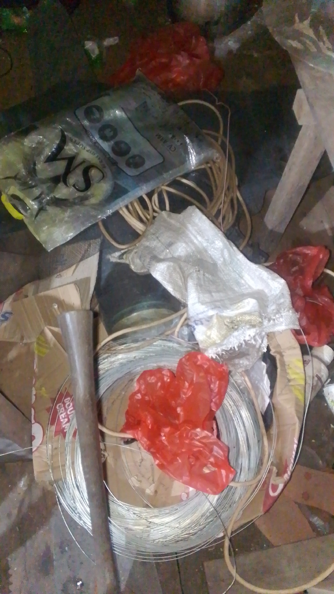 A close-up, overhead shot captures a chaotic pile of various materials in what appears to be an outdoor storage area or a cluttered workspace. The scene is dominated by spools of thin, silvery wire, some of which are covered by a crinkled, bright red plastic bag. A rough, cylindrical metal pipe lies across a large cardboard box, and another coiled bundle of yellowish-tan cord is visible in the background.

Among the clutter, a plastic bag with the "SM" logo and what appears to be electronic components or a remote control is partially visible. A crumpled white burlap-like sack sits atop some of the wire spools, and parts of other cardboard boxes are scattered around. The ground surface is uneven and appears to be dirt or concrete with debris. The overall impression is one of disorganization and accumulated supplies, possibly for construction or repair work. The context of Olak-Olak Kubu, Indonesia, suggests a rural or semi-urban setting where such materials might be stored.
