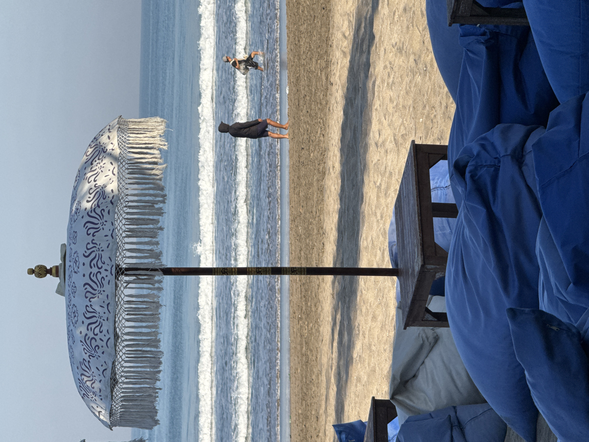 This image presents a sunny beach scene in Kuta, Indonesia, depicting a blend of cultural elements and leisure activities. The composition is horizontally divided into three main sections.
On the left, a traditional Balinese "Payung Tedung" style beach umbrella dominates the foreground. It features a white canopy adorned with intricate blue patterns and a fringe of white tassels. A golden finial caps its peak, and its dark pole displays a decorative band with unidentifiable symbols or patterns.
The central section captures the vast ocean and sandy shore. Multiple lines of white breaking waves stretch across the blue water, indicating a lively sea. Two people are visible in the ocean. Closer to the shore, one person dressed in a dark hooded top and dark shorts stands in the shallow water, facing away from the camera. Further out, another individual in lighter clothing appears to be engaged in activity within the waves. The sand is light brown, showing a transition from wet near the water's edge to drier further back.
On the right, several beach loungers or daybeds, covered in vibrant blue fabric cushions, suggest a comfortable relaxation area. Small wooden tables are positioned next to these seating arrangements. The clear, light blue sky above indicates bright, pleasant weather, typical for a beach day. The overall scene evokes a sense of tropical vacation and leisure at a popular Indonesian beach resort.