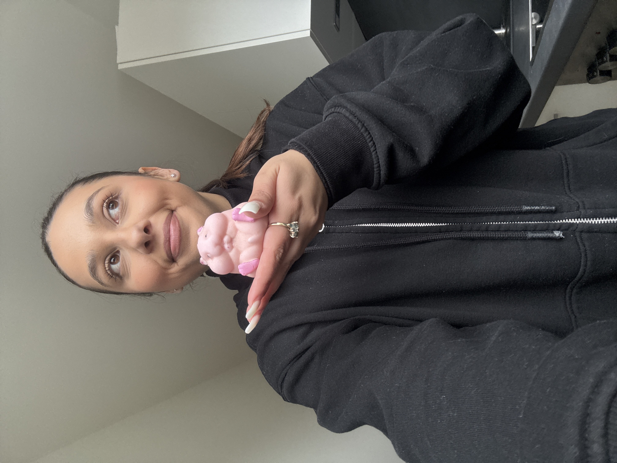 A woman with brown hair pulled back, wearing a black zip-up hoodie, is holding a small, pink, shaped object (possibly a toy or craft item) in her left hand. She is smiling slightly and looking upwards and to the left. A ring is visible on her left hand. The background includes a white wall and what appears to be a partial view of a cabinet or shelf. The photo was taken indoors. The location is not identifiable as being in Mönchengladbach, Germany, based on the provided image alone.
