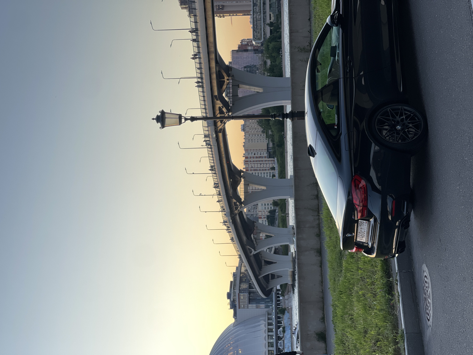 In a city setting, a black BMW M4 coupe is parked on the right side of an asphalt road. The car is positioned facing away from the viewer, with its rear end closer to the camera. The vehicle's license plate, visible on the rear bumper, reads "У460СТ 198". To the left of the car is a strip of green grass and a concrete barrier. Beyond the barrier, a body of water is visible, with a modern bridge spanning across it in the background. The bridge is supported by large white pillars and has a walkway with people strolling along it. On the far left of the image, a large, domed building with white architectural features is partially visible. The sky above is a soft gradient of light blue and yellow, indicating either sunrise or sunset. A vintage-style lamppost stands in the mid-ground, to the left of the bridge. The scene depicts an urban environment with a focus on the parked car and the scenic backdrop of the bridge and cityscape.