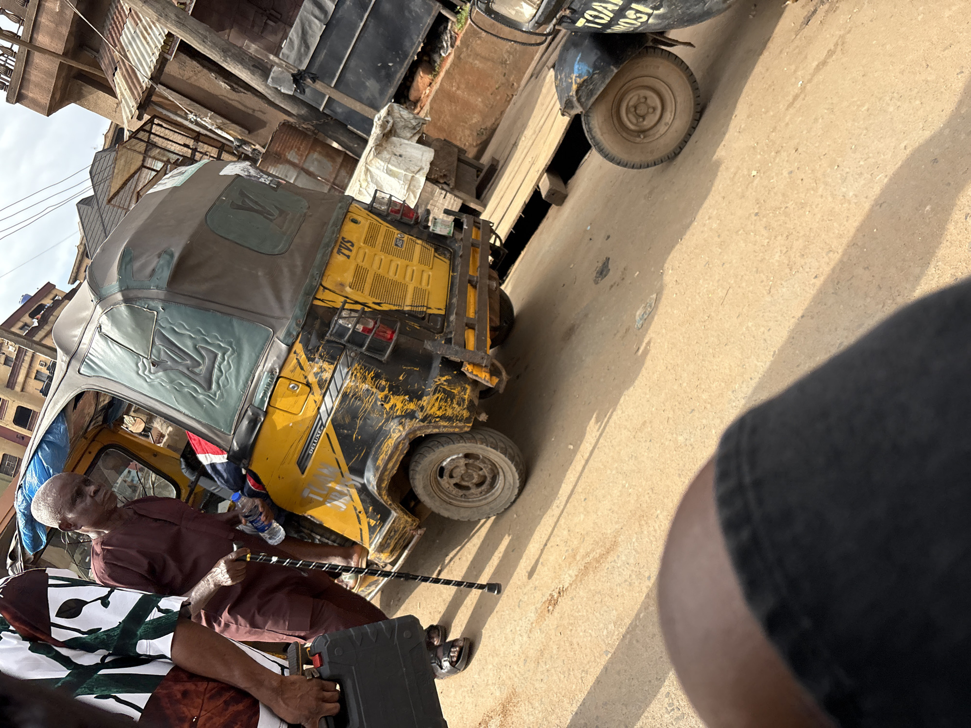 The image depicts a street scene in Onitsha, Nigeria, focusing on a yellow auto-rickshaw, also known as a "Keke Napep." An elderly man, dressed in a brown traditional outfit, is standing next to the auto-rickshaw, holding a walking stick and a bottle of water. He appears to be disembarking or preparing to embark. To his left, another person, partially visible, is holding a black case. The auto-rickshaw has a weathered appearance, with visible scratches and wear on its yellow body. The roof of the auto-rickshaw is covered in a brown material with a Louis Vuitton pattern printed on it. The background reveals the urban environment of Onitsha, with buildings constructed from corrugated metal and concrete, suggesting a bustling commercial area. The ground is paved with compacted dirt or sand. Another auto-rickshaw is partially visible in the foreground on the right side of the image.