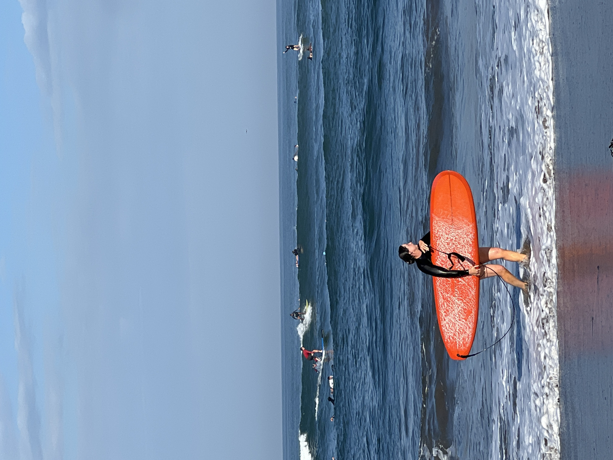 In North Kuta, Indonesia, on a beach, a woman in a black wetsuit walks out of the water carrying a bright orange surfboard. Her legs are submerged in the shallow, foamy water. In the background, several other individuals are surfing on the blue ocean waves under a clear, pale blue sky with sparse clouds. Some surfers are standing on their boards, while others are in the water.