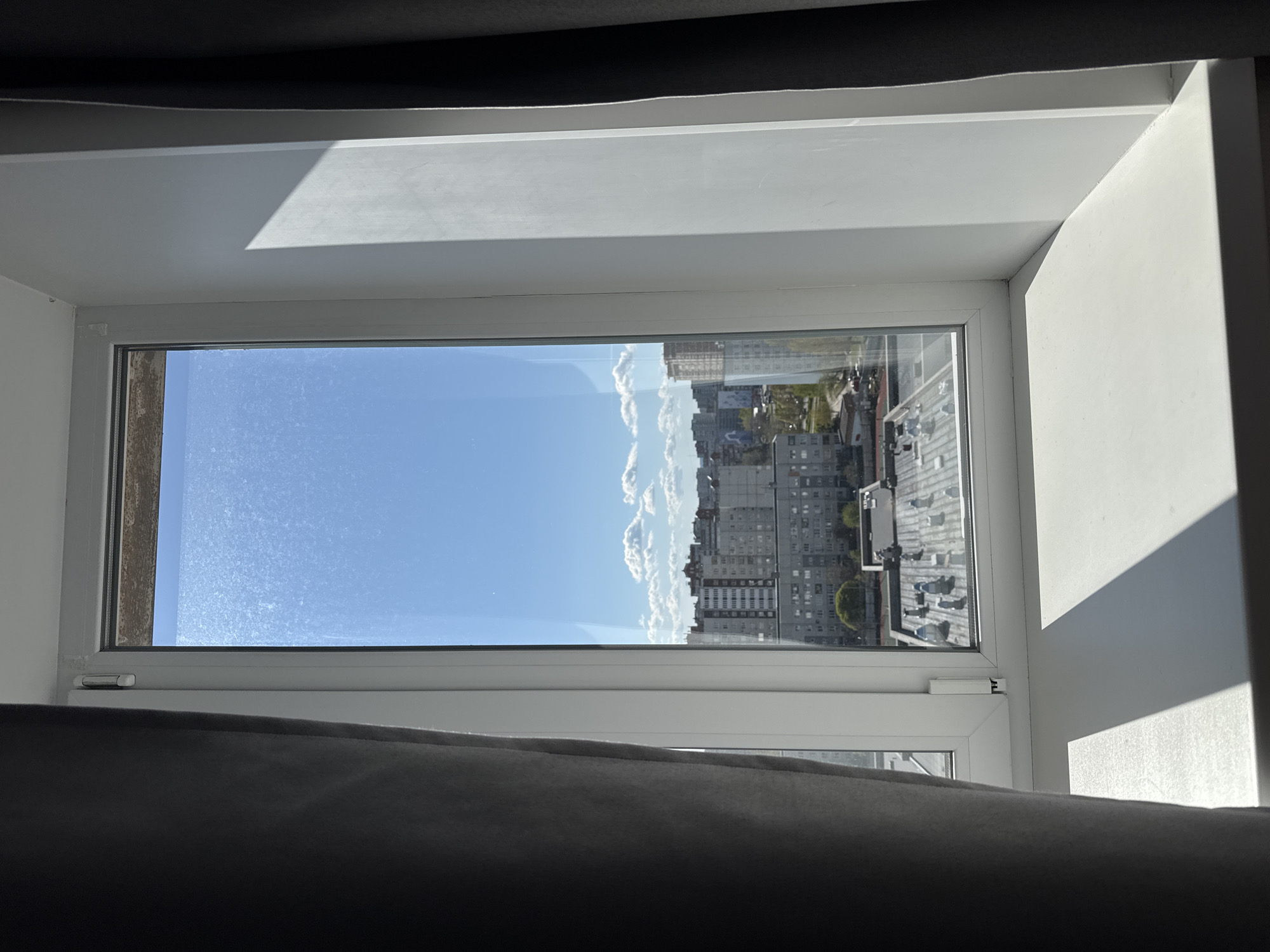 The image, if rotated clockwise 90 degrees, shows a bright daytime view from indoors, looking out through a white-framed window onto an urban cityscape. The white window frame and sill are prominent, with a dark, possibly fabric, material partially visible at the top and bottom, suggesting curtains or blinds. The window glass appears somewhat dirty or dusty.
Beyond the window, a clear blue sky with scattered white clouds occupies the upper portion of the view. Below the horizon, a dense array of multi-story buildings, typical of a city, stretches across the landscape. These structures are predominantly grey or light-colored, featuring numerous windows and balconies.
Bright sunlight illuminates the scene, casting sharp, angular shadows on the white window frame, sill, and the adjacent wall on the right, indicating a clear, sunny day. No people are visible, and no specific activity is taking place. There is no discernible text in the image.