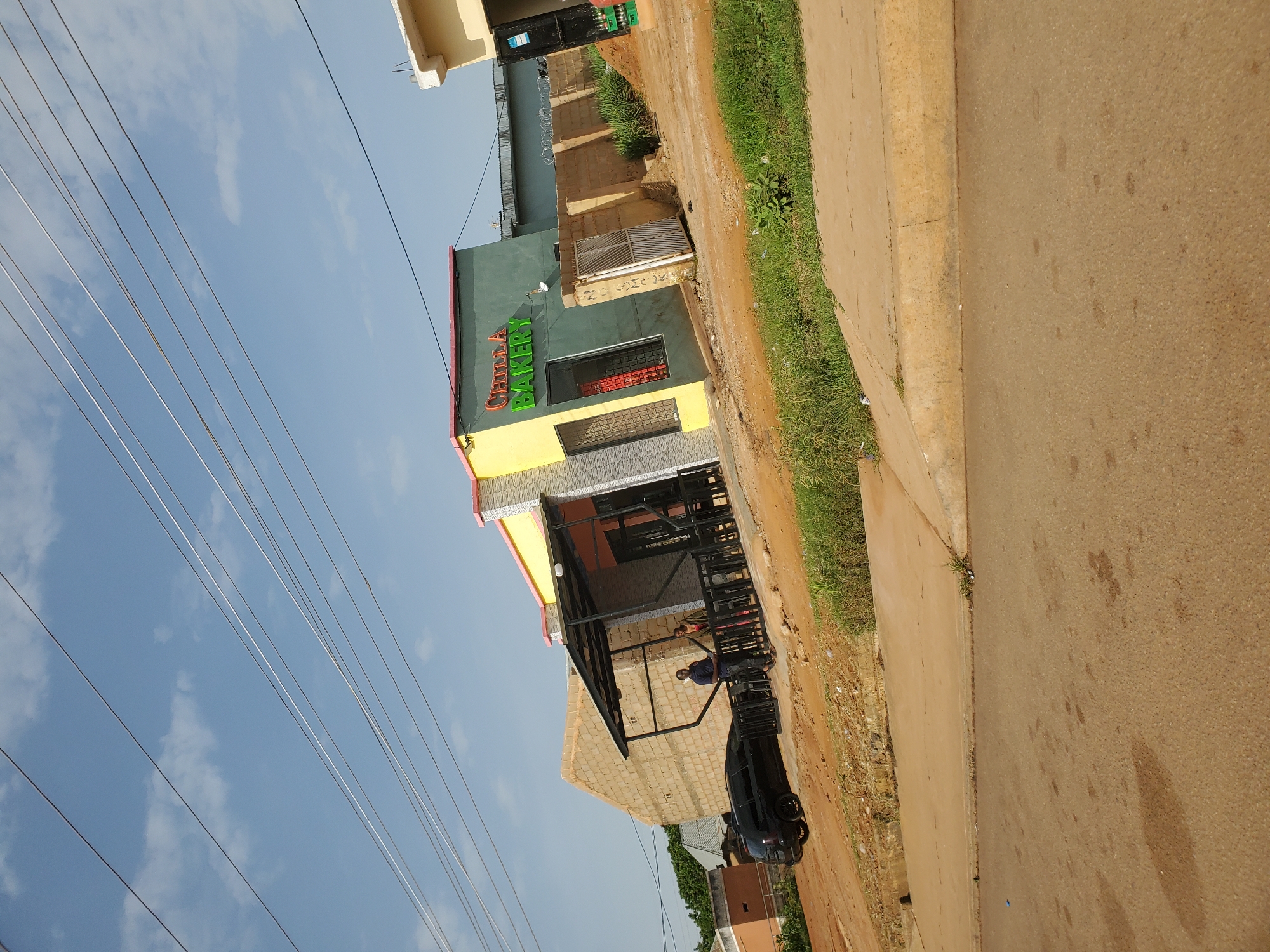 In Bukuru, Nigeria, an image captures the exterior of a building identified as "CHILLA BAKERY" with green lettering against a teal facade. The bakery features a yellow front with a pink trim along the roofline. A covered outdoor seating area with black railings and a metal roof is positioned in front of the bakery, accessible from a dirt and gravel area. Two individuals are standing near the seating area, one man in a blue shirt and jeans, and another person whose gender is not clearly discernible. A black SUV is parked to the left of the bakery. Behind the bakery, construction is evident with a brick wall. The scene is illuminated by daylight under a sky with scattered clouds. A paved road runs in the foreground.