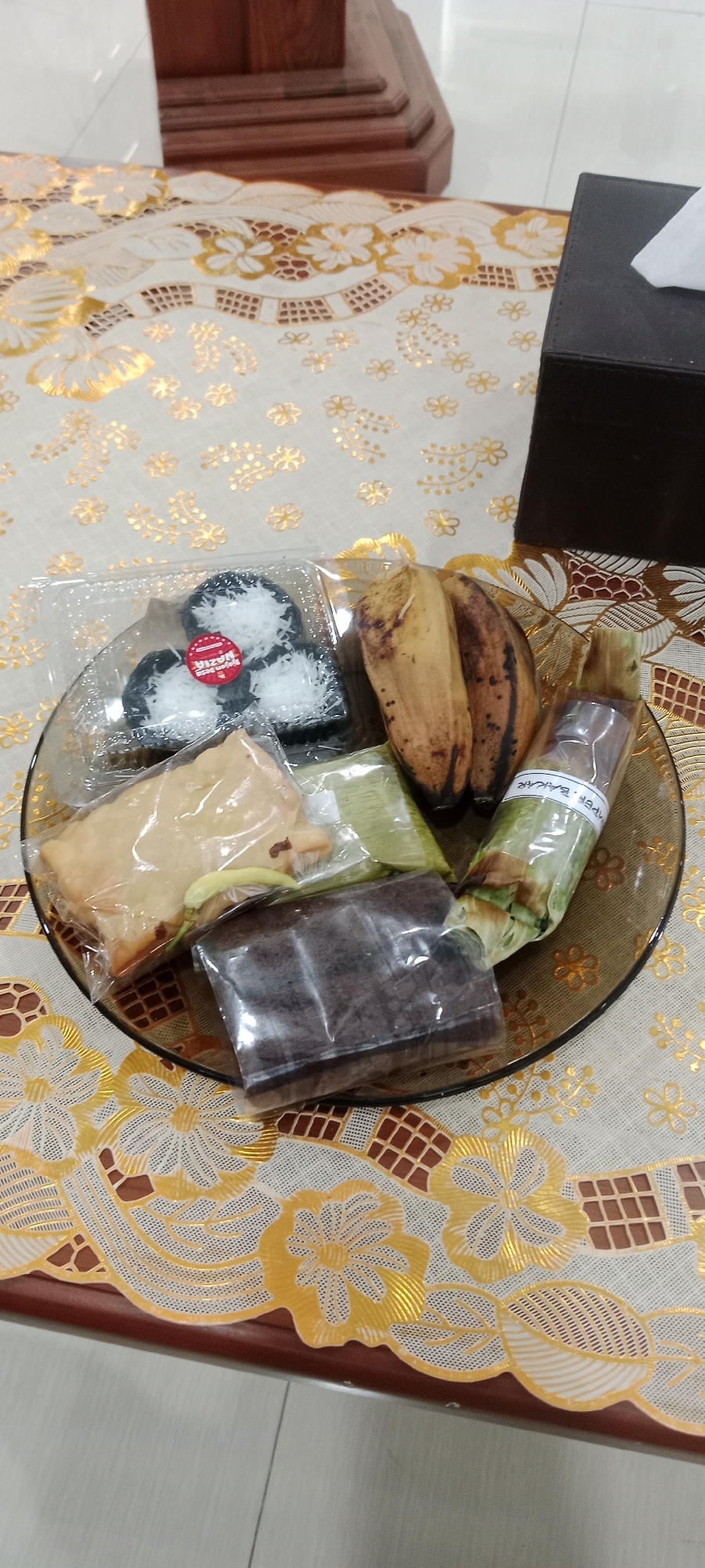 A clear, overhead shot displays a round, dark brown glass plate filled with various food items, resting on a white tablecloth with intricate gold floral patterns and a brown checkered border. The plate contains a small pile of three dark gray, circular pastries topped with shredded white coconut, each sealed in clear plastic. Adjacent to these are two overripe bananas with brown spots, and further to the right, a long, thin item wrapped in green banana leaves and clear plastic, with text that appears to read "MPER BAKAR". On the lower part of the plate, there's a light brown, rectangular pastry wrapped in plastic, and below it, a dark brown, rectangular pastry also in plastic. A small green chili pepper is visible peeking out from under the light brown pastry. In the background, a dark brown tissue box with a white tissue peeking out sits to the right. The tablecloth is set on a dark wooden table, partially visible at the bottom of the frame, and beyond that, a light-colored tiled floor. The scene is indoors, likely on a table in Tuban, Indonesia, given the context of the food items which appear to be local snacks.