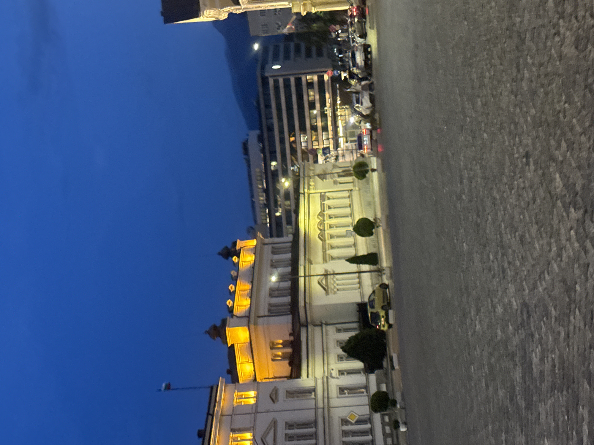 The image depicts an urban scene at twilight in the city of Sofia, Bulgaria. The sky is a deep blue with faint cloud formations. In the foreground, a large, dark grey paved area, resembling a cobblestone square or plaza, occupies the lower half of the frame.
The mid-ground features several buildings. To the left, a multi-story white building with neoclassical architectural elements is prominently illuminated in warm yellow light on its upper floors and roofline. A flagpole atop this building displays the white, green, and red stripes of the Bulgarian flag. Adjacent to it and slightly further back, another white building of similar style is also brightly illuminated in yellow. In front of these buildings, a street is visible with several parked cars, including a distinct yellow compact car. Various streetlights are active, casting light on the scene.
Further back, a modern, multi-story building with horizontal window bands is visible, also illuminated. Beyond this, a dark mountain range (Vitosha mountain) rises against the twilight sky. A single bright light source, possibly the moon or a strong streetlight, is visible above the mountain. On the street, multiple vehicles are present, some with visible headlights and taillights, suggesting ongoing traffic. Sparse trees and shrubs are planted along the street and near the buildings. Due to the low light and distance, individuals are not distinctly identifiable, but indistinct figures are perceptible in the mid-ground near the street.