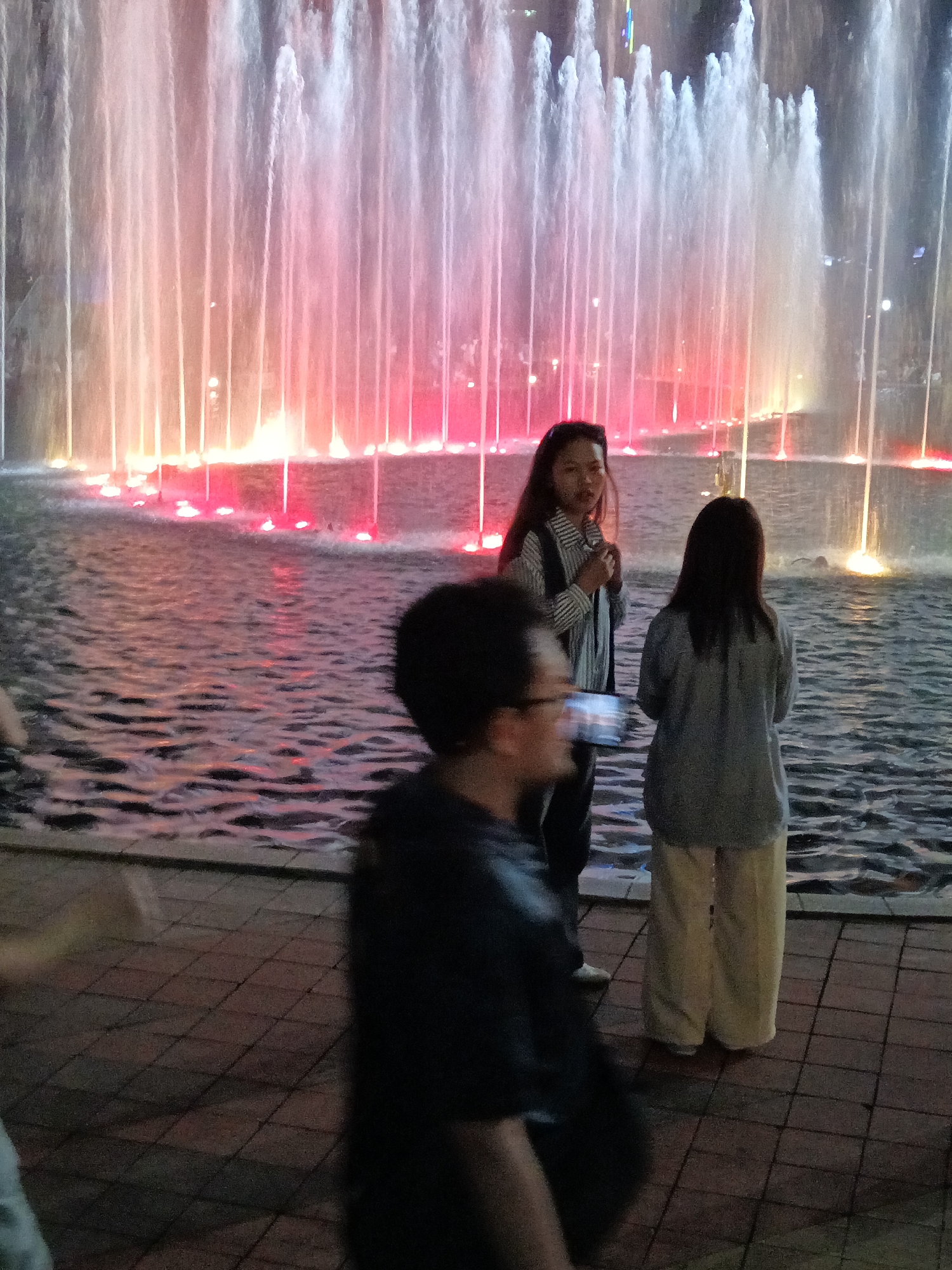 A group of people stand by a fountain in the city of Kuala Lumpur, Malaysia. The fountain is illuminated with colorful lights and shoots water into the air, creating a dramatic display. In the foreground, a person with glasses is seen from behind, holding a phone. To their right, a woman in a striped shirt is looking towards the fountain. Behind her, another woman in a gray sweater stands with her back to the camera. The ground is paved with tiles, and the water of the fountain creates ripples. The scene appears to be at dusk or night, given the lighting.