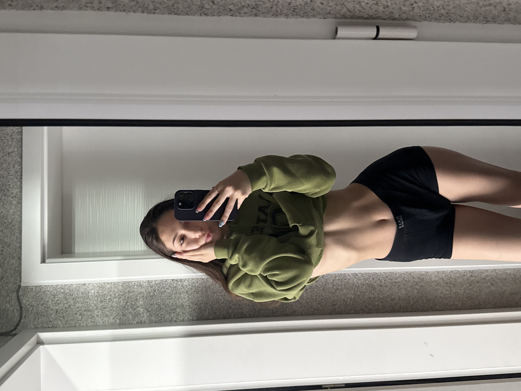 A mirror selfie taken indoors captures a young person, likely in Sofia, Bulgaria. The subject, a person with long, dark hair, stands with their mid-torso visible. They are wearing an olive-green, cropped long-sleeve top that reveals their abdomen, and black shorts or athletic underwear.
The person holds a dark-colored smartphone, equipped with multiple camera lenses, in their left hand to capture the reflection. Their fingers, adorned with light blue nail polish, are visible on the phone. Their right hand is gently resting on their cheek, partially obscuring their face, while they look towards the camera with a relaxed expression.
The background reflected in the mirror shows an interior space. A white door frame or wall molding is prominent, along with a plain white wall. Below, a light grey, speckled carpet or floor covering is visible. On the black shorts, white text can be seen, appearing upside down, which reads "XXAM OMOU". The lighting in the scene is bright and even, suggesting either natural daylight or well-lit artificial conditions.