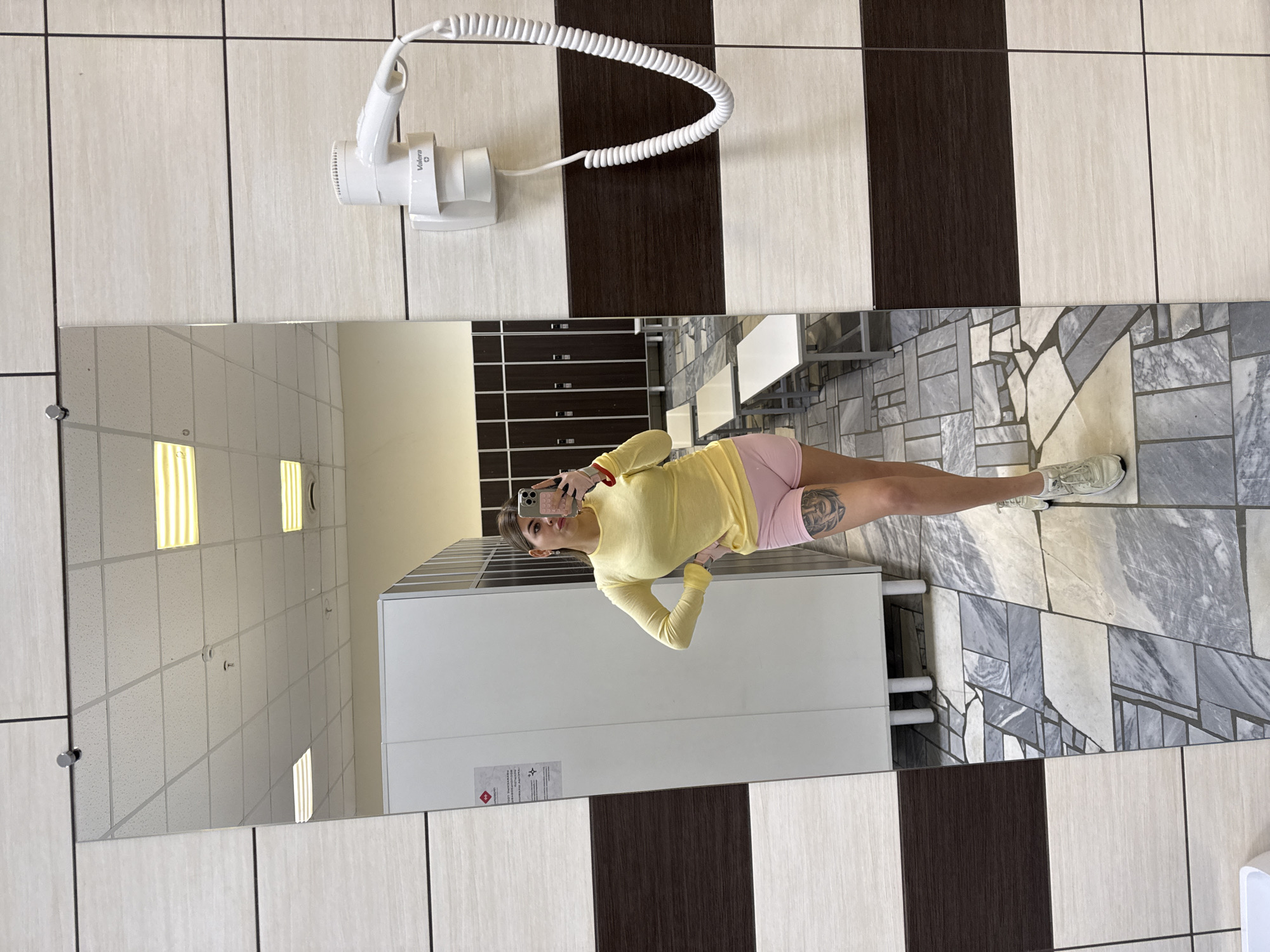 A woman is taking a mirror selfie in what appears to be a public restroom, potentially in a fitness center or similar facility, located in Chaikovsky, Russia. The woman is wearing a yellow long-sleeved shirt and pink shorts, and her phone is visible in her hand. Her reflection is visible in a long, horizontal mirror spanning the width of the image. The restroom has tiled walls with alternating light and dark brown vertical stripes and a tiled floor with a gray mosaic pattern. A wall-mounted hairdryer is visible on the wall above the mirror. Lockers and possibly other restroom fixtures are visible in the mirror's reflection.