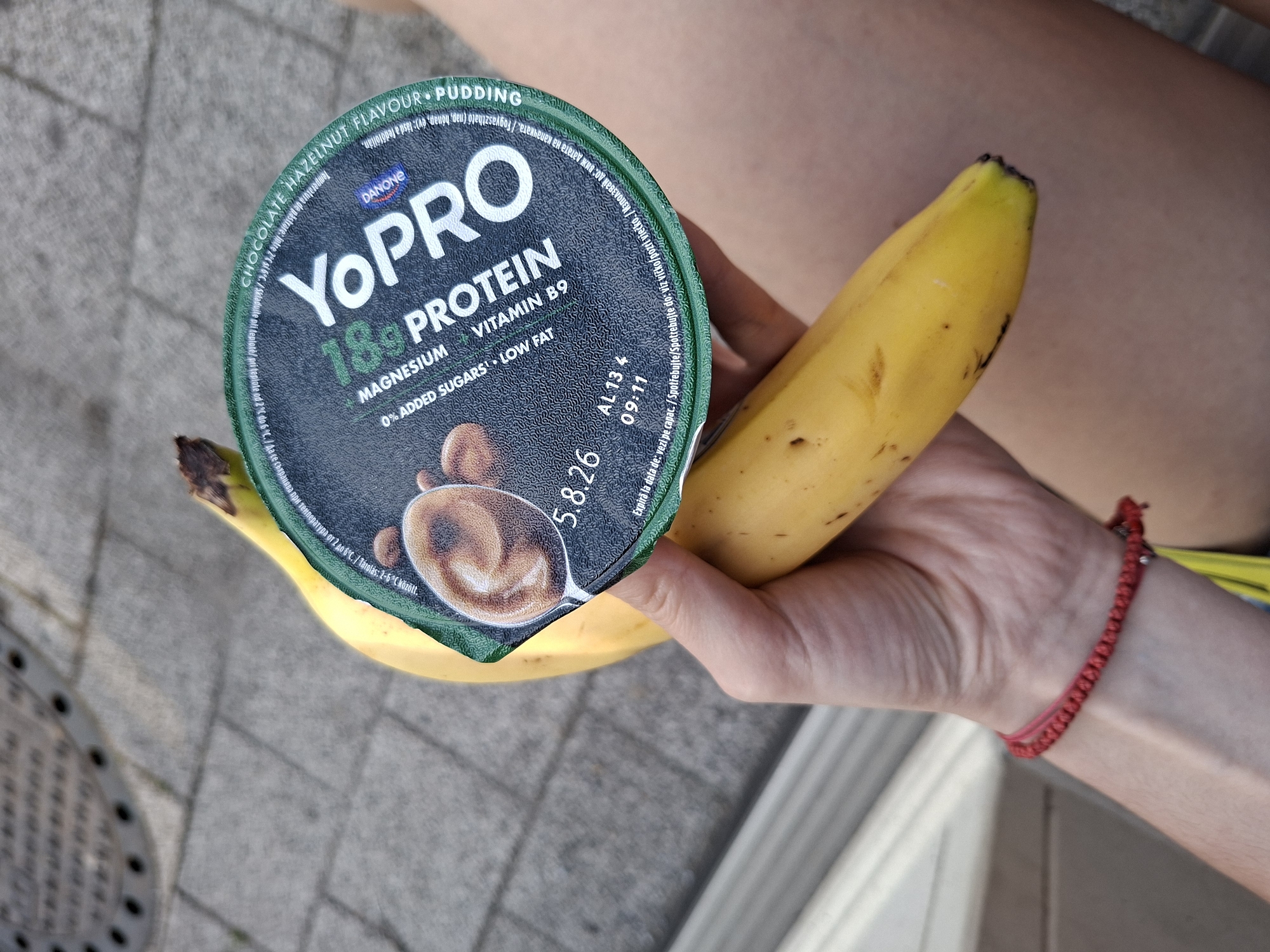 The image captures a first-person perspective of a person's hand holding a snack outdoors during the daytime. The main subjects are a yellow banana and a dark green Danone YoPro protein pudding cup, held against the person's leg.
The person's hand, visible from the wrist downwards, holds both items. A red braided bracelet is worn on their wrist. The background shows grey pavement tiles, with a portion of a metal grate visible on the left, suggesting an urban or outdoor setting. The light appears natural, indicating it's not nighttime.
The YoPro pudding cup is prominently displayed, showing its lid. Key details on the lid include "CHOCOLATE HAZELNUT FLAVOUR PUDDING," "18g PROTEIN," "MAGNESIUM," "VITAMIN B9," "0% ADDED SUGARS," and "LOW FAT." The brand "DANONE YOPRO" is clearly visible. The best-before date "5.8.26" is printed on the lid, along with instructions in multiple languages, including "Temperatura de pastrare: intre 2°C si 6°C." (Storage temperature: between 2°C and 6°C), and phrases like "Expiră la data de: vezi pe capac." (Romanian for "Expires on: see lid") and "Използв" (part of "Използвайте" - "use" in Bulgarian), confirming the multi-lingual packaging often found in the Balkan region, consistent with the Plovdiv, Bulgaria context. Other text includes "AL 13 4" and "09:11," likely production or batch codes. The image on the lid depicts a spoonful of chocolate hazelnut pudding.
The banana appears ripe, with some dark spots on its skin. The overall scene suggests a casual snack moment.