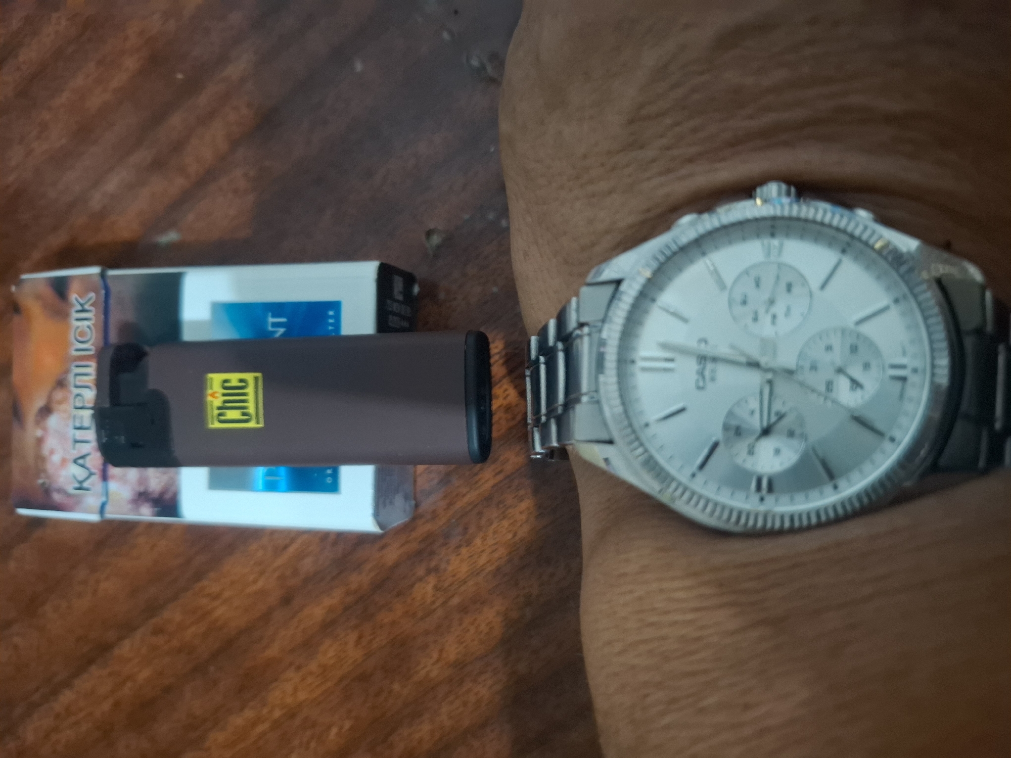 The image is a top-down shot of a wooden table with a pack of "Chic" cigarettes and a brown lighter on it. A person's wrist wearing a silver wristwatch is visible in the foreground. The watch face shows multiple subdials and Roman numerals. The background shows a grainy wooden surface. There is no indication of the city of Almaty, Kazakhstan.