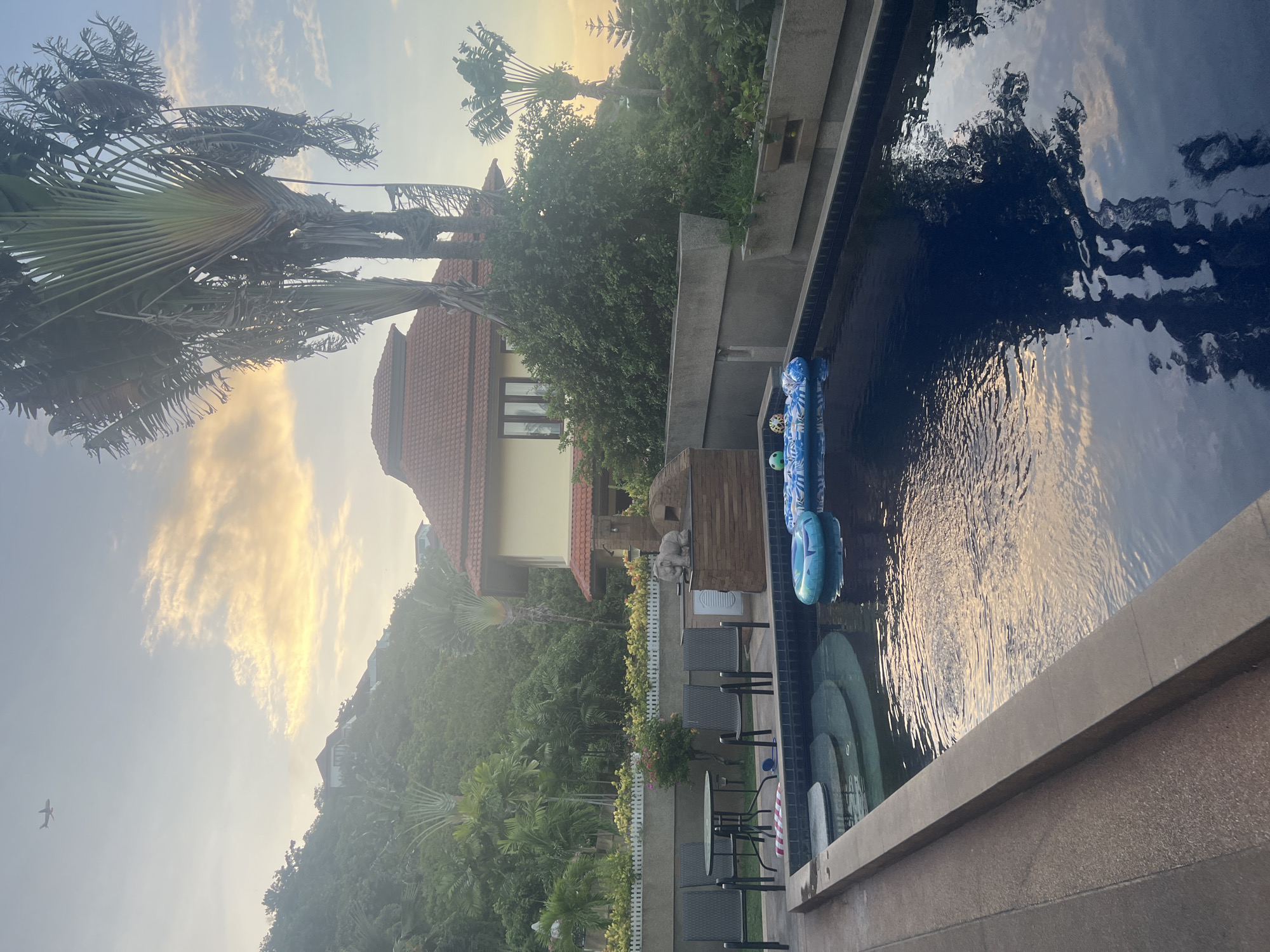 An outdoor swimming pool area is depicted in Ko Samui District, Thailand, bathed in the soft light of dusk. The pool, filled with dark blue water, has a stepped entry area on the left. Two inflatable pool floats are visible in the water, one is blue with white patterns and the other is primarily blue with a white design. The poolside is paved with textured concrete. A low, beige-colored wall with a concrete coping runs along the right side of the pool. To the left of the pool, a small round table with four chairs is set up. A small white picket fence with yellow flowers behind it lines a raised garden bed further back.
Behind the pool area, a multi-story house with a red tile roof and yellow walls is situated on a lush, green hillside. Several large, palm-like trees with fan-shaped leaves are prominent in the landscape, as are numerous other trees and dense foliage. The sky above is a pale blue, with wispy clouds tinged with yellow and orange, indicating sunset. An airplane can be seen flying in the upper left corner of the image. A stone sculpture of an elephant stands on a stone structure to the right of the table and chairs. The overall impression is one of a serene, tropical residence.
