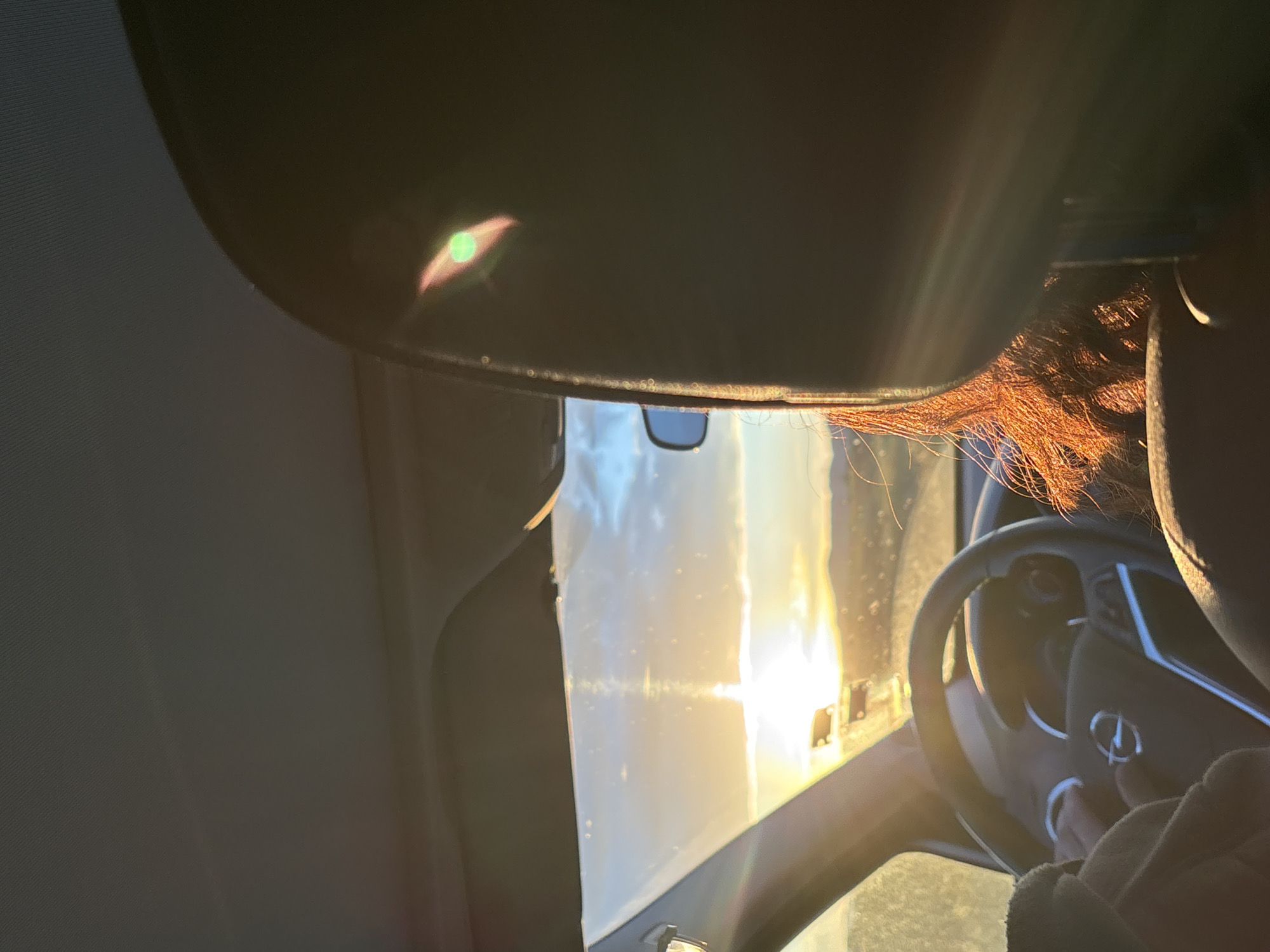The image, taken from the interior of a vehicle, shows a driver obscured by the sun visor and bright sunlight through the windshield. The steering wheel is visible, with a hand partially visible on the lower right. Reddish-brown hair is visible near the visor. The outside scene is blurred by the sun and the angle. The location is unclear as there are no distinct landmarks. The city name is irrelevant without visual confirmation.