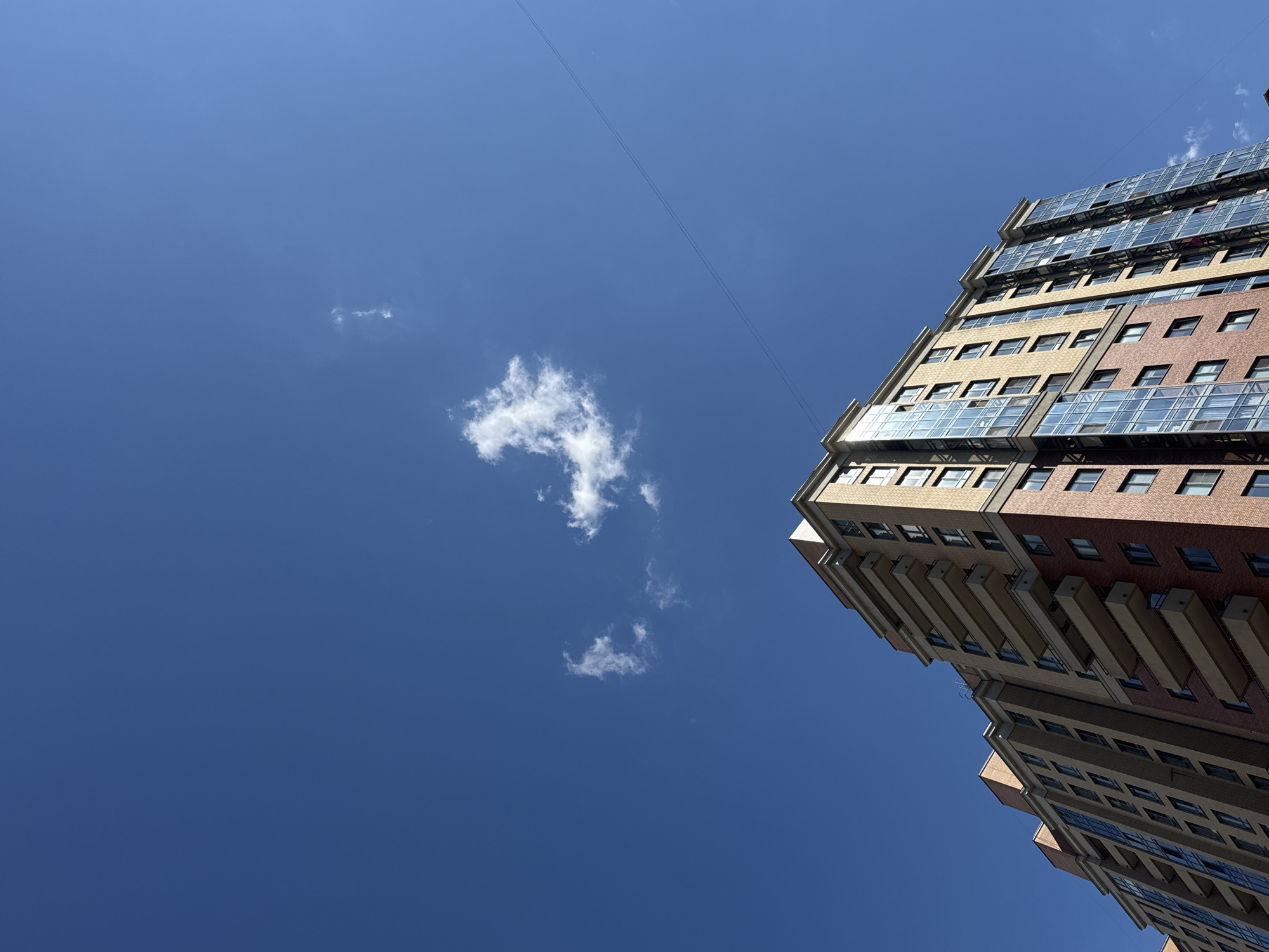A low-angle, wide shot captures a modern, multi-story apartment building against a clear, bright blue sky with a few wispy clouds and a thin wire stretching diagonally across the upper portion of the frame. The building, constructed with a combination of beige and reddish-brown brick, features numerous windows and balconies. The architecture suggests it is an apartment complex in Kudrovo, Russia. The sun casts shadows on the building's facade, indicating it is daytime. There are no people visible in the frame. The environment appears to be urban.