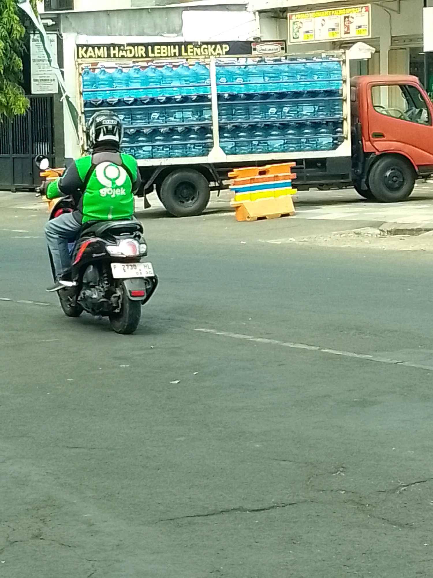 A Gojek delivery driver wearing a green jacket and helmet is riding a scooter on a road in Jember, Indonesia. The driver is positioned in the foreground, with the scooter moving away from the camera. In the background, a large truck is parked, loaded with many large blue water bottles. The truck has text in Indonesian that reads "KAMI HADIR LEBIH LENGKAP" which translates to "WE ARE HERE MORE COMPLETE". There are also colorful traffic cones stacked on the side of the road. The scene appears to be a street in a commercial or industrial area, with buildings and trees visible in the distance. The road surface is asphalt, with some cracks and debris present. The lighting suggests it is daytime.