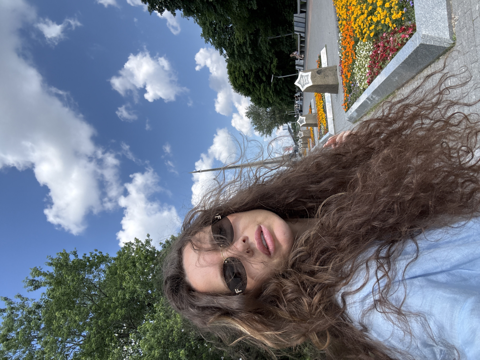 The image shows a woman with long, curly brown hair and sunglasses, wearing a light blue shirt. She is taking a selfie in what appears to be a park or public square. In the background, there are flowerbeds with colorful flowers, stone monuments, benches, trees, and a blue sky with white clouds. There are also other people in the background, some sitting on benches and others walking. The setting suggests a bright, sunny day. The specific location in Moscow is likely the "Victory Park" (Park Pobedy) on Poklonnaya Hill, known for its monuments, flower arrangements, and expansive green spaces.