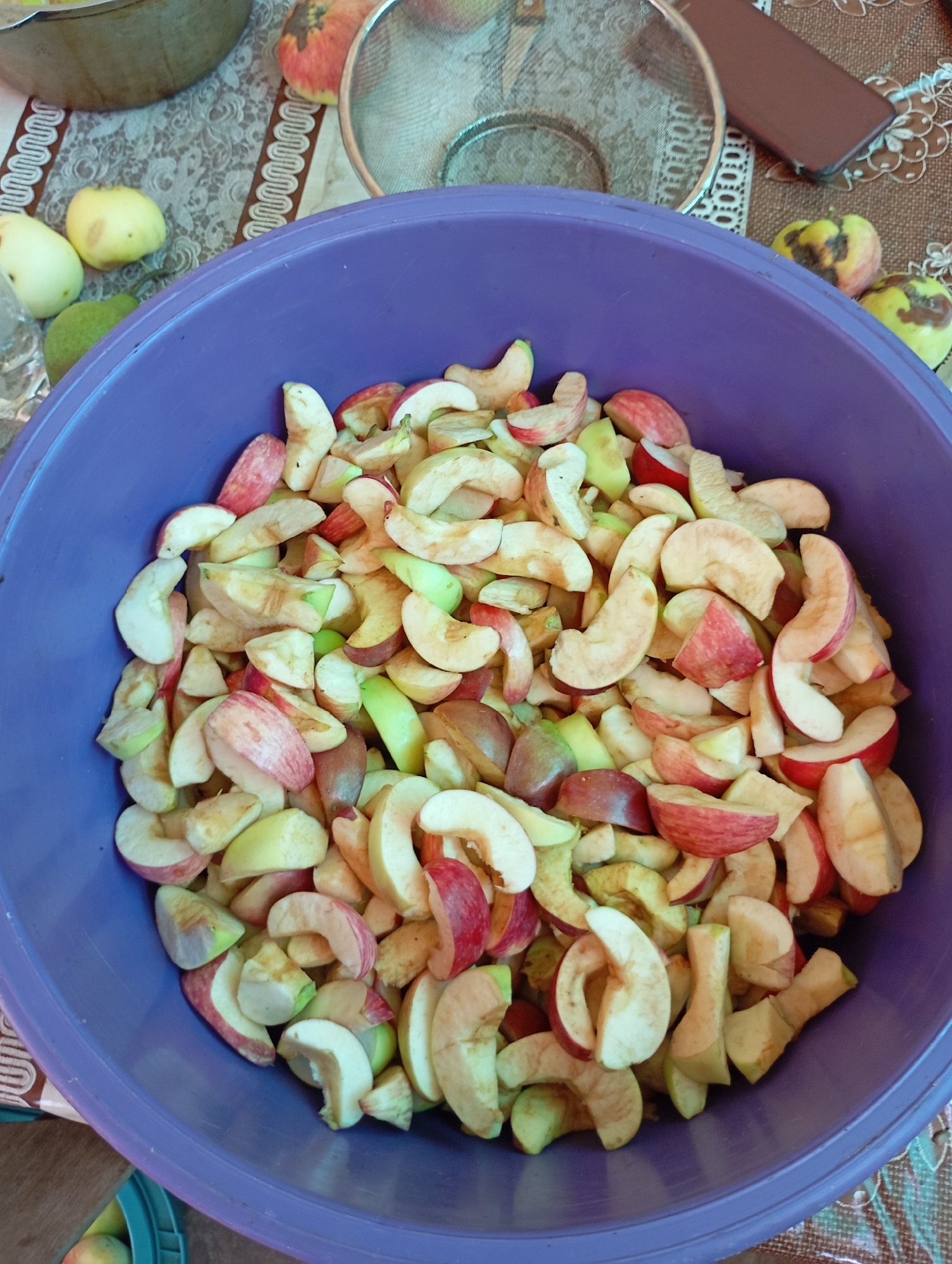 In Horishni Plavni, Ukraine, a large purple bowl is filled with chopped apples. The apples are sliced into wedges and pieces, with visible skin and flesh. Some pieces show red skin, while others are primarily yellow or green. The bowl sits on a patterned tablecloth, which has a border of white and brown decorative elements. To the left of the bowl, a few whole apples and pears are visible on the tablecloth. In the background, a metal sieve and a dark object, possibly a phone, are also present on the tablecloth. The scene appears to be indoors, likely in a kitchen or dining area, given the presence of food preparation items.