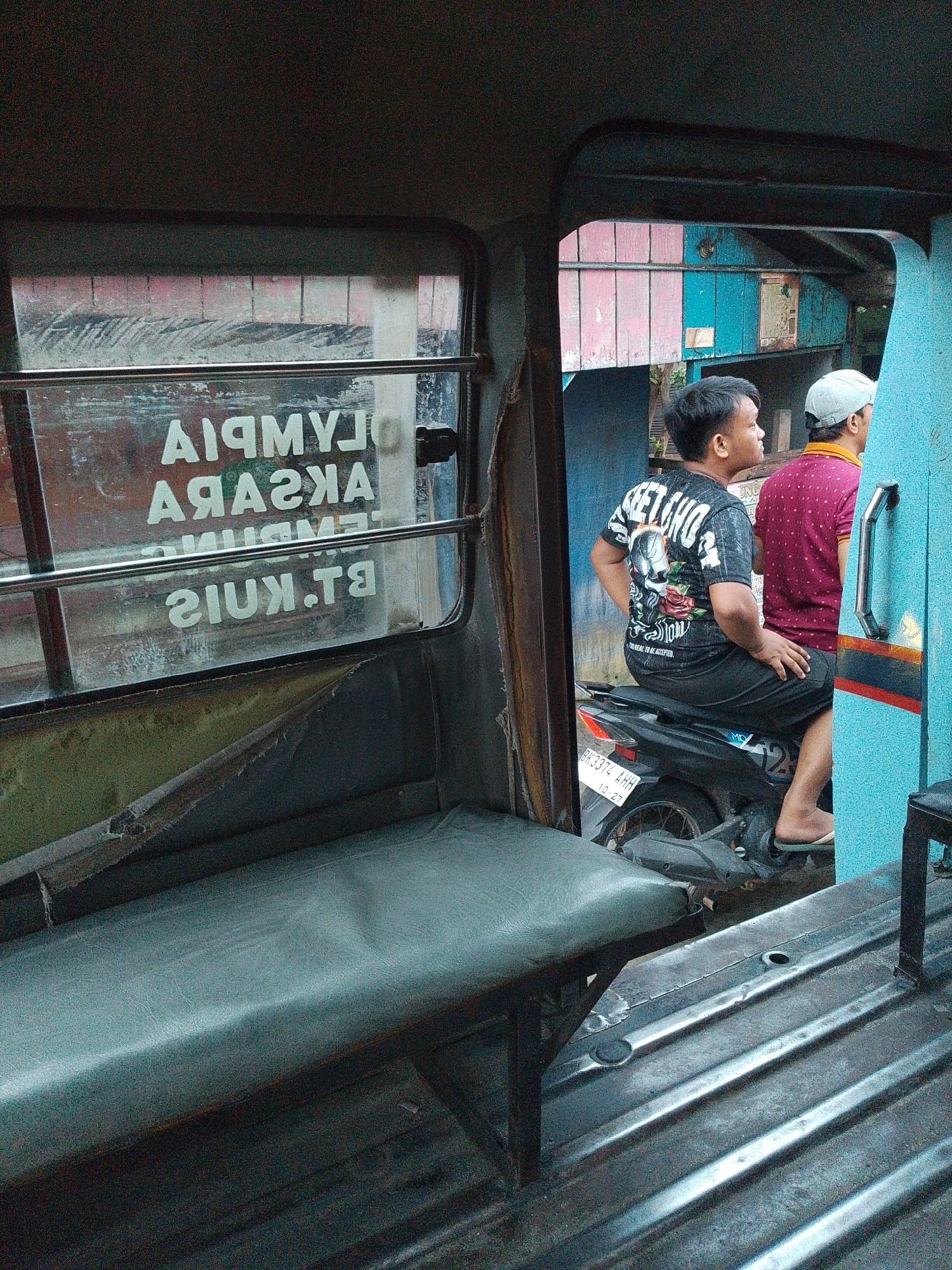 The image depicts the interior of a vehicle, possibly a type of public transport, in Bandar Khalipah, Indonesia. Two young men are visible. One, wearing a black t-shirt with a skull graphic and black shorts, is seated on a black motorcycle parked behind the passenger compartment. The other, wearing a maroon shirt and a white cap, stands next to him. Reflected in the glass window of the vehicle, text in white capital letters appears reversed, reading "OLYMPUS" and "AKASRA". The interior features worn, grey upholstered seats and a ribbed metal floor. The exterior visible through the window suggests an outdoor environment with a building featuring pink and blue colored panels. The license plate on the motorcycle reads "BK3374 AH".