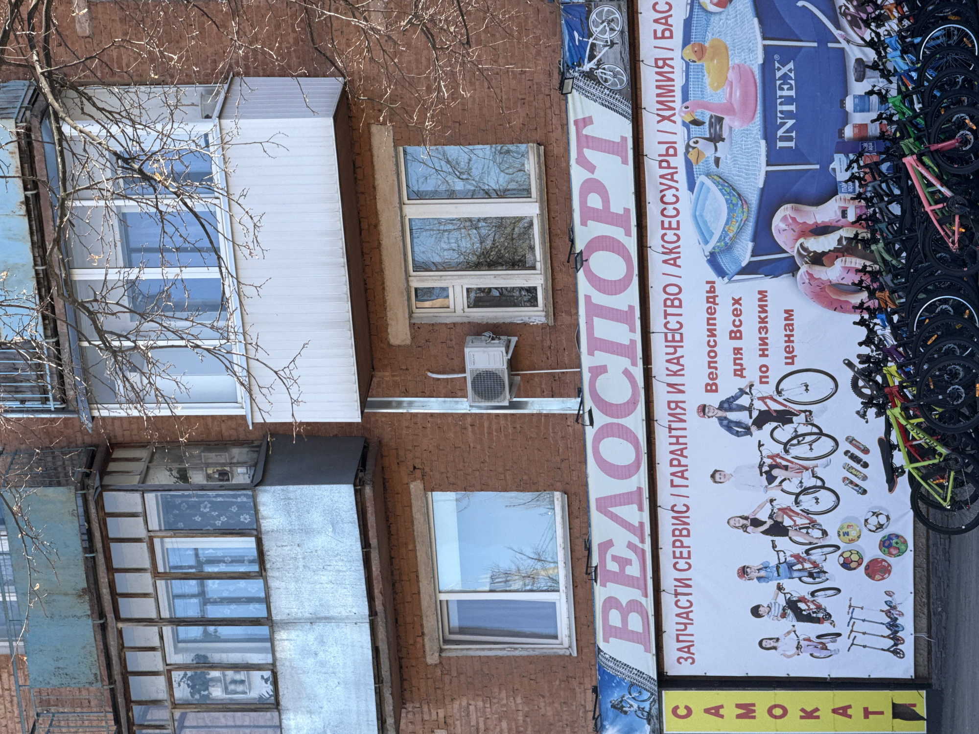 The image depicts the exterior of a brick building, likely in Votkinsk, Russia, indicated by the Cyrillic text on a sign. The sign promotes "VELOSPORT" (cyclesport) and lists services: parts, service/warranty and quality, accessories, chemistry/basins (likely for swimming pools), and also includes text with a bicycle advertisement. The building has windows and a balcony structure, one of which is enclosed with metal sheeting. Bicycles are stacked to the side of the advertisement.