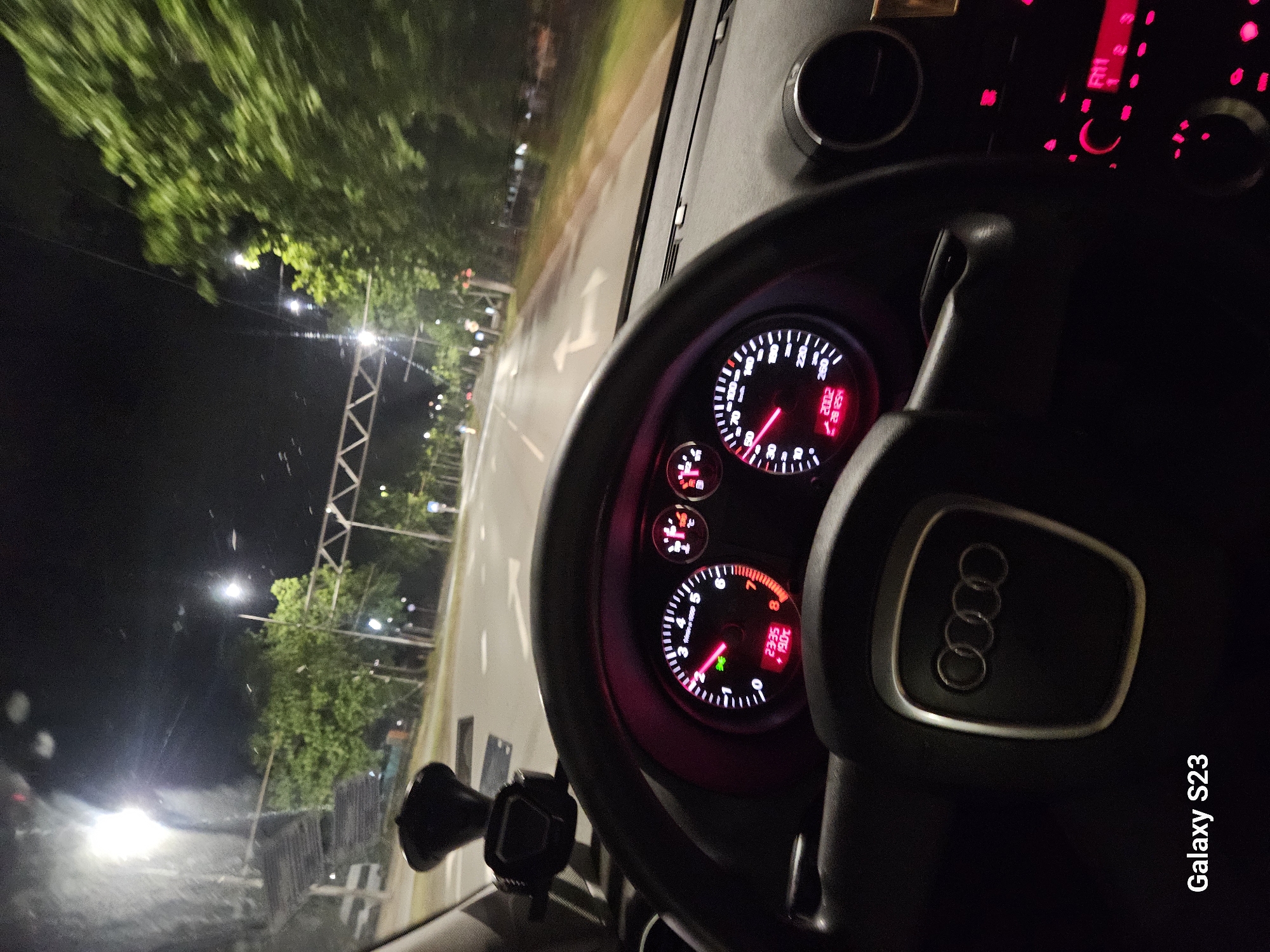 The image shows the interior of an Audi car at night, likely in motion. The dashboard is visible, displaying illuminated gauges. The steering wheel is prominently featured with the Audi logo. Outside, the scene suggests a road with trees and streetlights, potentially within the city of Sliven, Bulgaria. There are no people visible in the image. A phone mount is attached to the dashboard. The words "Galaxy S23" are visible in the lower right corner, likely indicating the device used to take the picture.