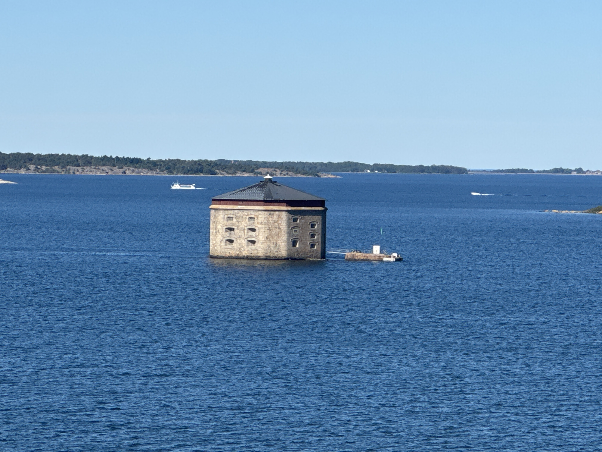 The image depicts a cylindrical, stone fort situated in the blue waters of the Baltic Sea, in the archipelago of Karlskrona, Sweden. The fort, which appears to be a historical defensive structure, has a dark, conical roof and several small, arched windows visible on its exterior. A small, connected platform with a white structure and what appears to be a small boat is attached to one side of the fort. The water is calm with gentle ripples. In the background, a long, tree-covered island coastline stretches across the horizon under a clear blue sky. Several small boats and a larger ferry-like vessel are visible in the distance, navigating the sea. The overall scene is peaceful and evokes a sense of historical significance and natural beauty.