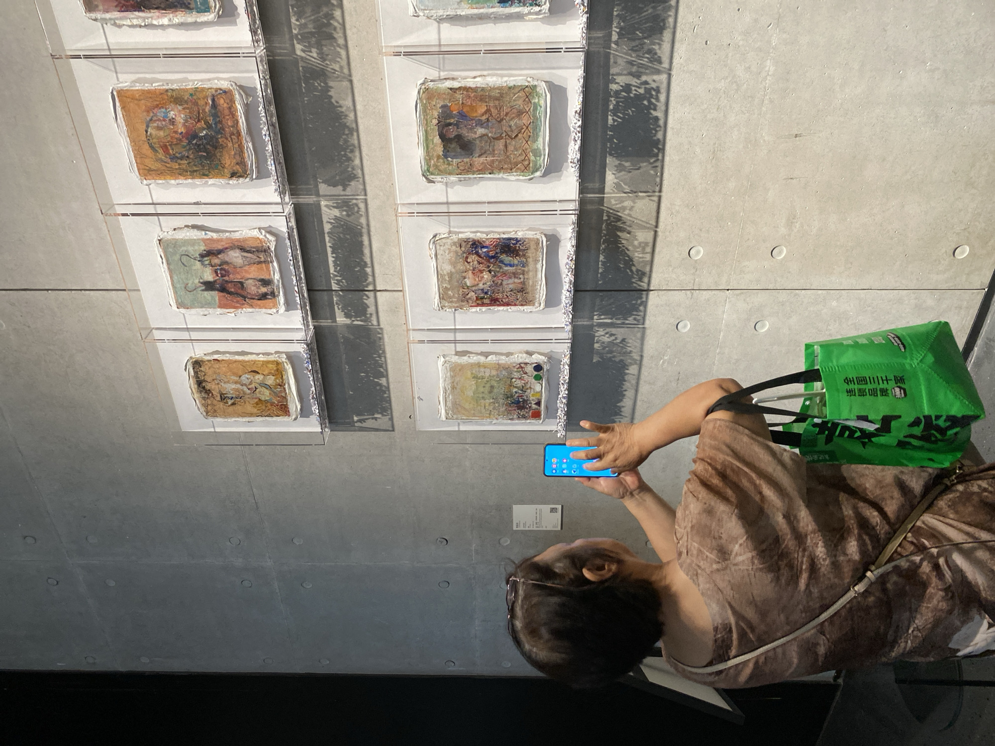 An elderly East Asian woman, viewed from behind, stands in front of a concrete wall in an art gallery. She is wearing a brown patterned t-shirt and glasses perched on her head. The woman is holding a smartphone in her right hand, aiming it at artworks displayed on the wall. Her left hand is also near the phone, possibly assisting with the camera. A green reusable shopping bag hangs from her left arm. The artworks appear to be small, framed pieces with textured, abstract imagery. The wall is constructed of large, grey concrete panels with circular indentations. Shadows from above cast a patterned effect on the wall and artworks. The overall environment suggests a modern, minimalist art exhibition space.