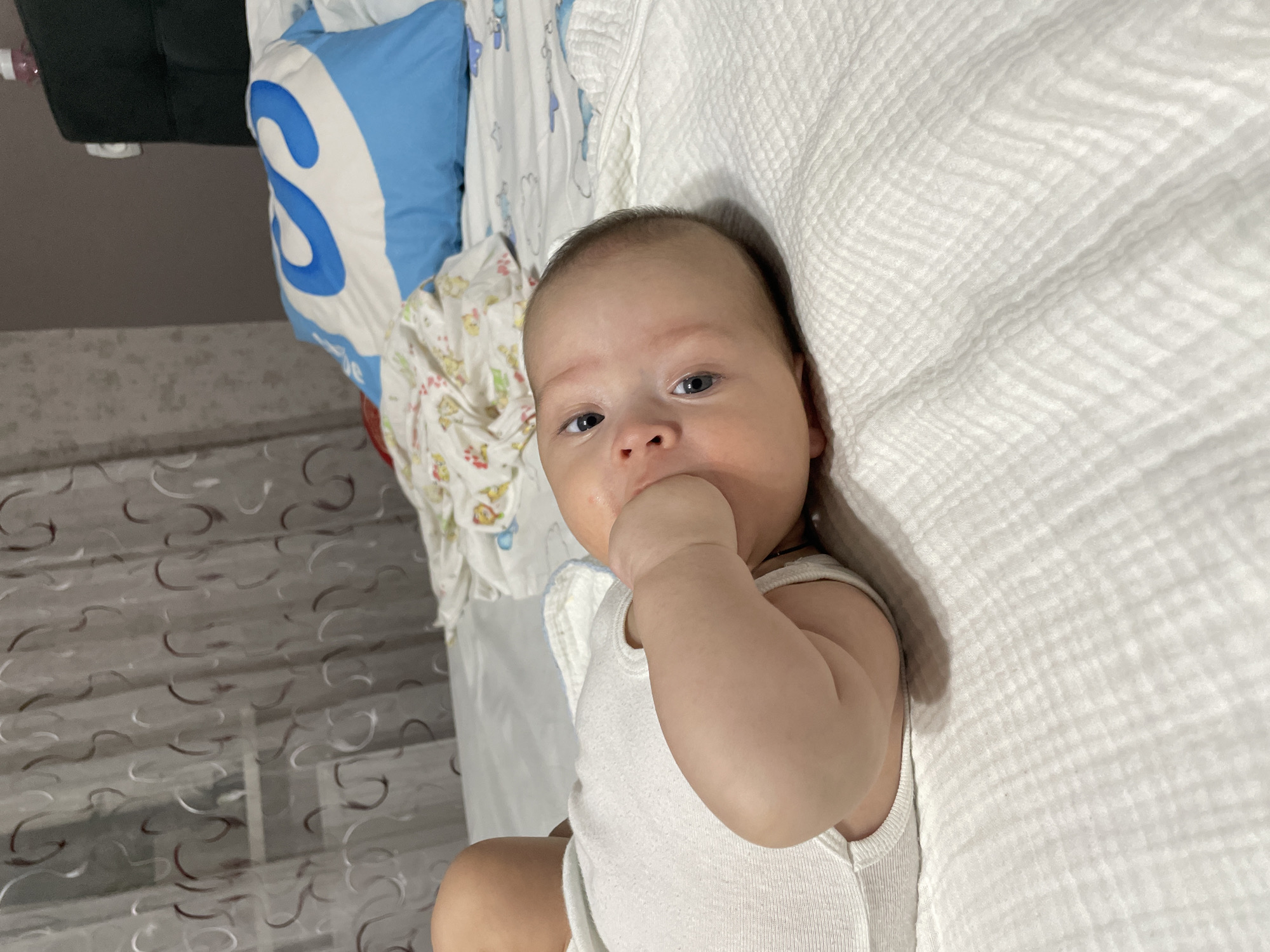 A newborn infant lies on a white, textured bedsheet, with its right hand in its mouth. The infant is wearing a white onesie. Behind the infant, a blue and white pillow with a large "S" logo is visible, along with a patterned blanket. The background includes a curtain with a swirling pattern and a dark headboard. The scene appears to be indoors, likely in a bedroom. The presence of a pillow and blanket suggests a domestic setting. There is no information in the image to determine its location within Fălești, Moldova.
